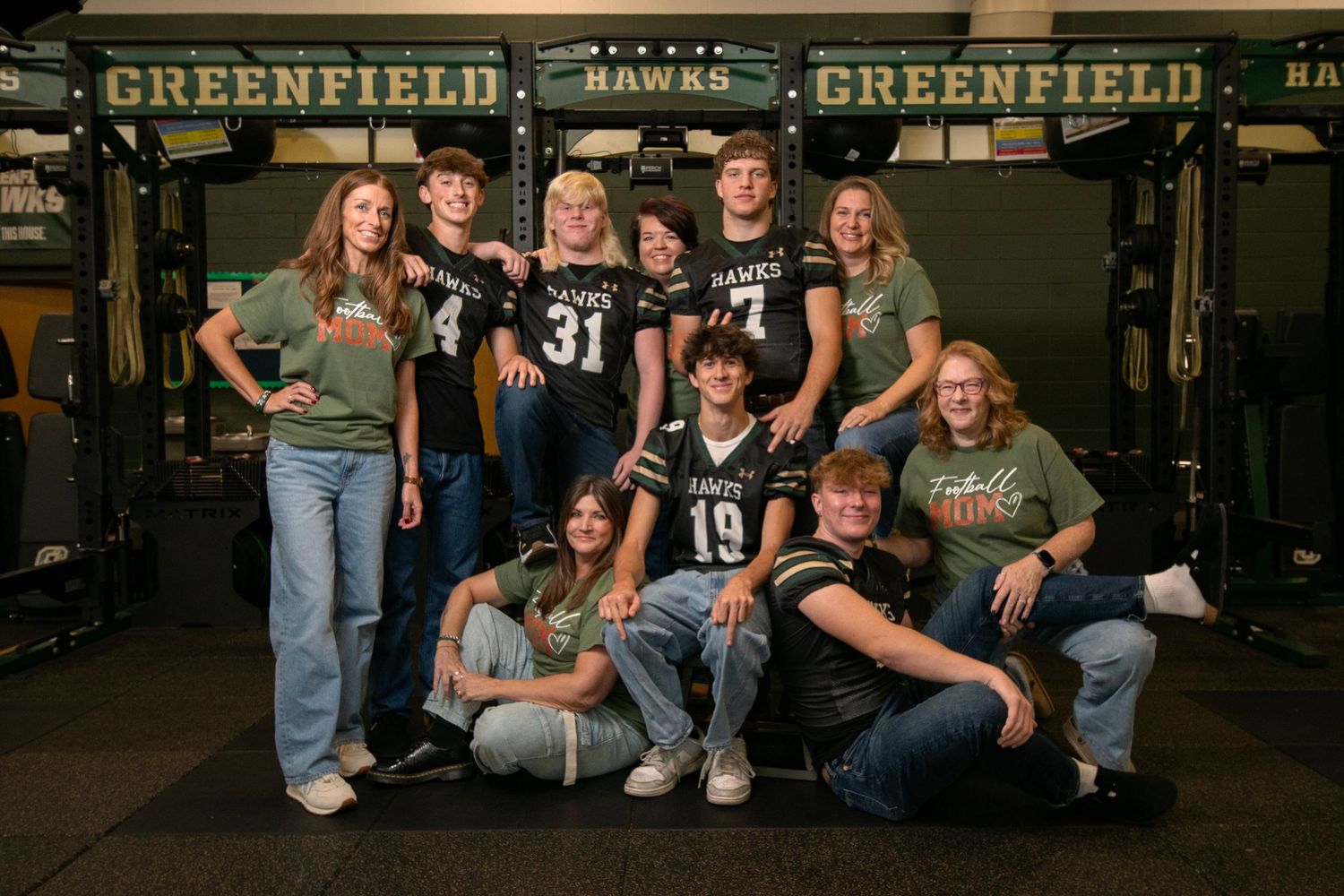 A group of athletes and supporters pose together in a high school weight room with Greenfield banners visible in background.