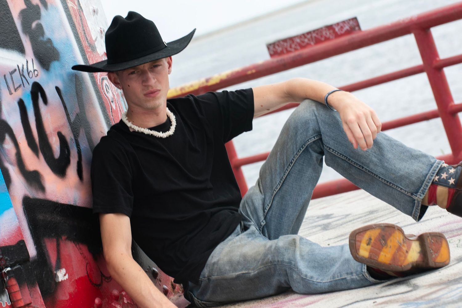 Young man in black cowboy hat and t-shirt with jeans sitting relaxed against colorful graffiti wall outdoors.