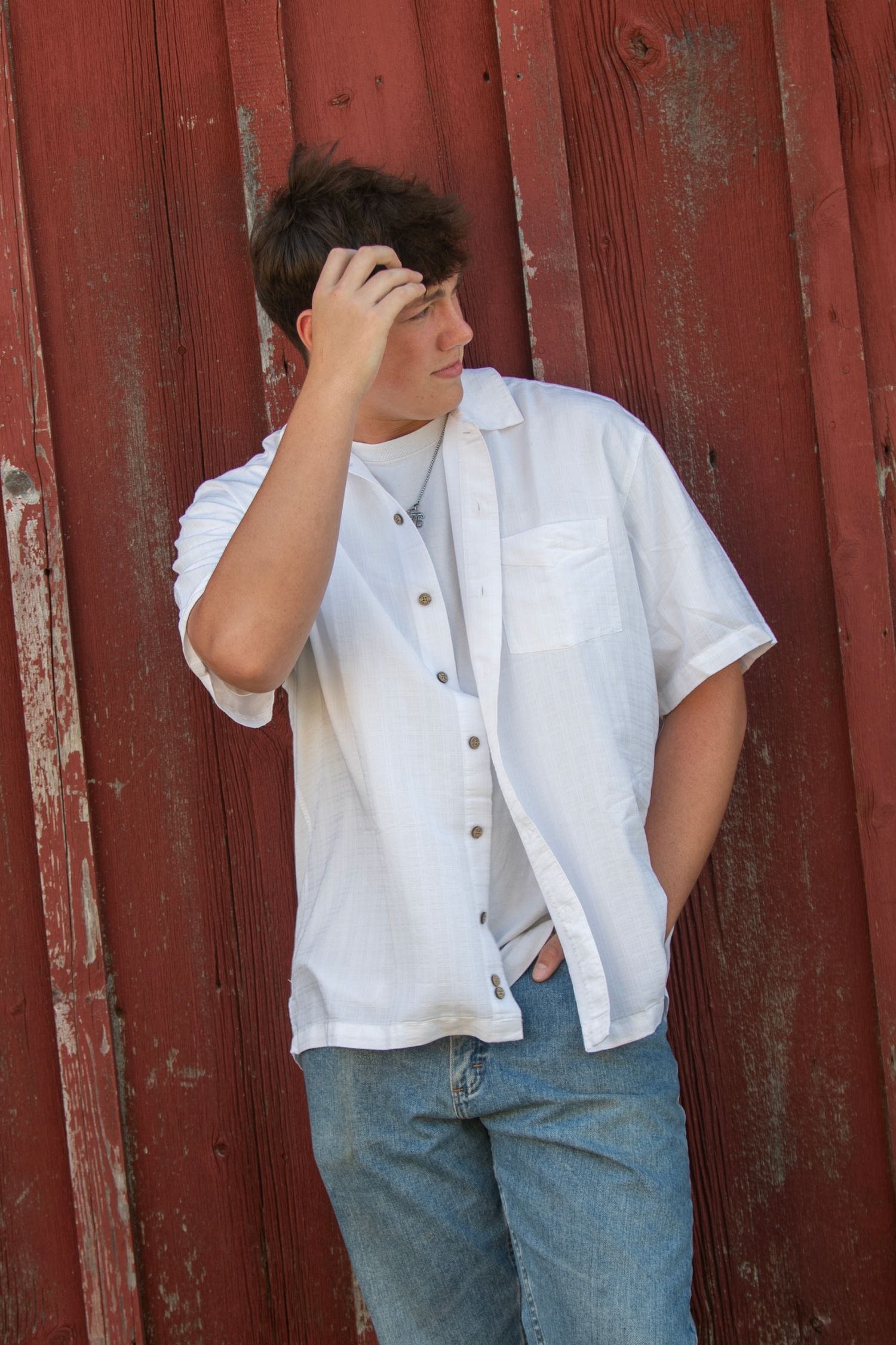 Teen boy in white shirt and jeans leaning against a red barn wall with hand raised to his head.