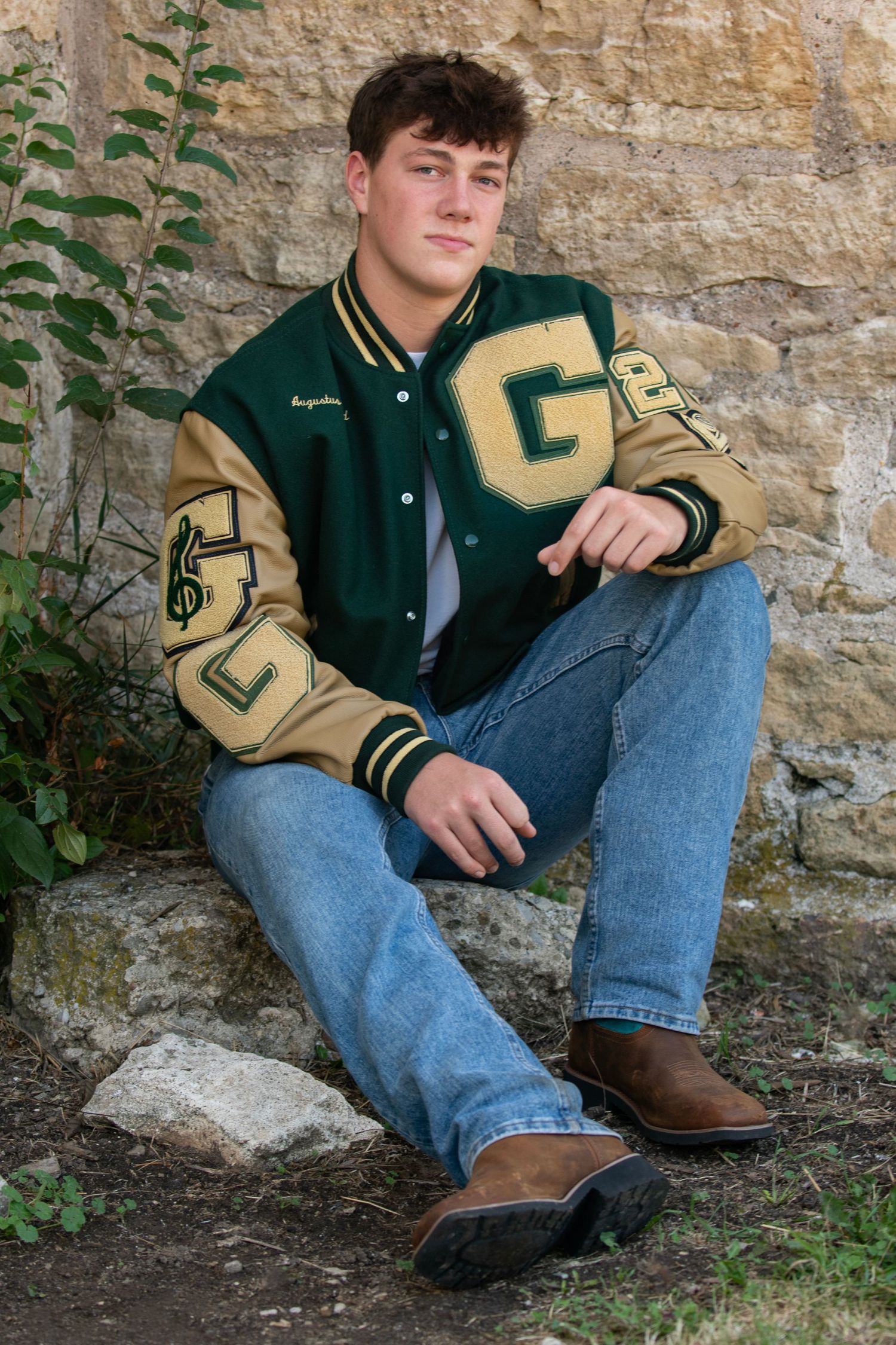 High school student wearing a green and gold varsity letter jacket sits casually against a stone wall.