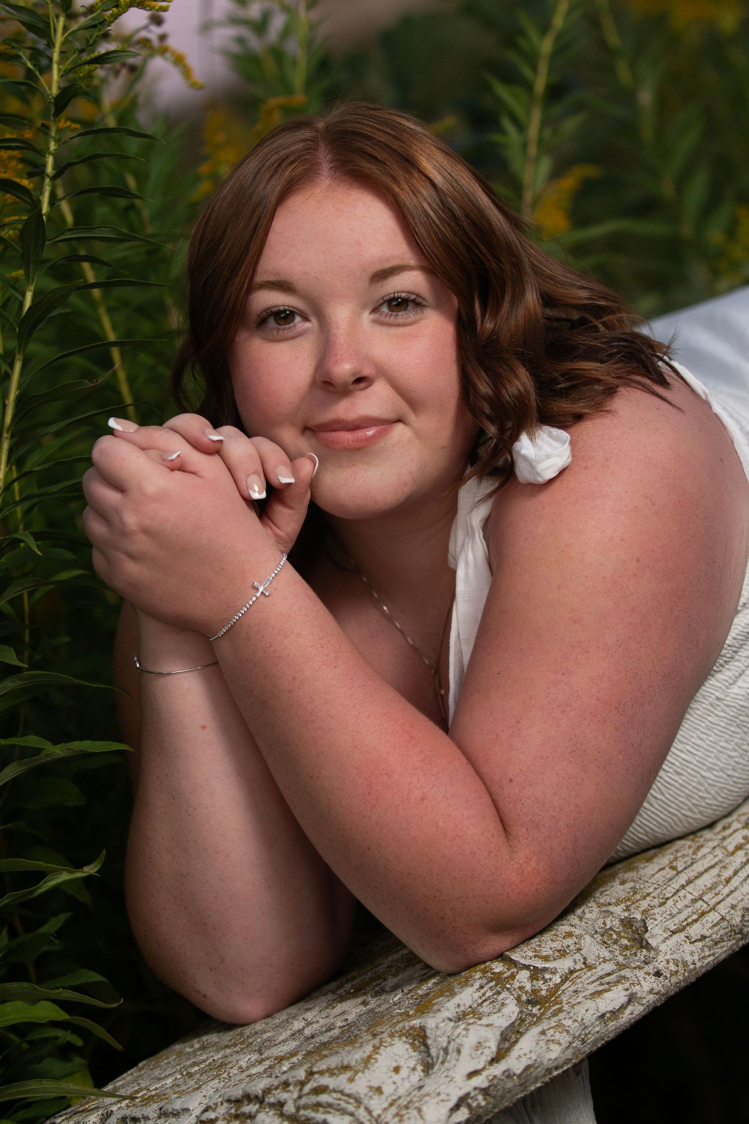 Young redhead woman in white dress leaning on rustic bench surrounded by yellow wildflowers smiling.