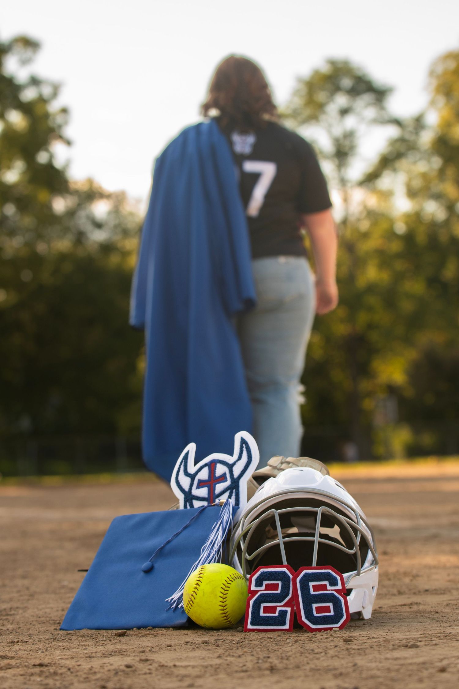 Softball helmet numbered 26, yellow ball, and graduation cap on dirt field with player in jersey number 7 holding blue gown.