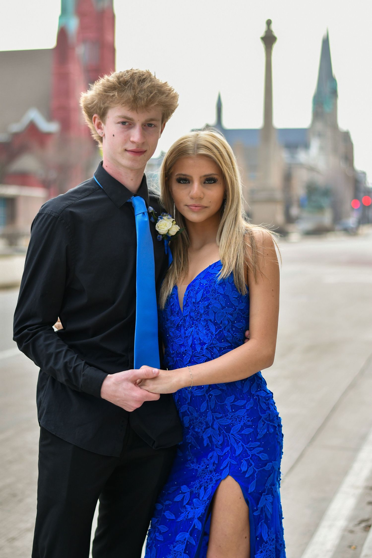 A teenage couple dressed in prom attire, the boy in black suit with blue tie and girl in blue gown, posing on a city street.