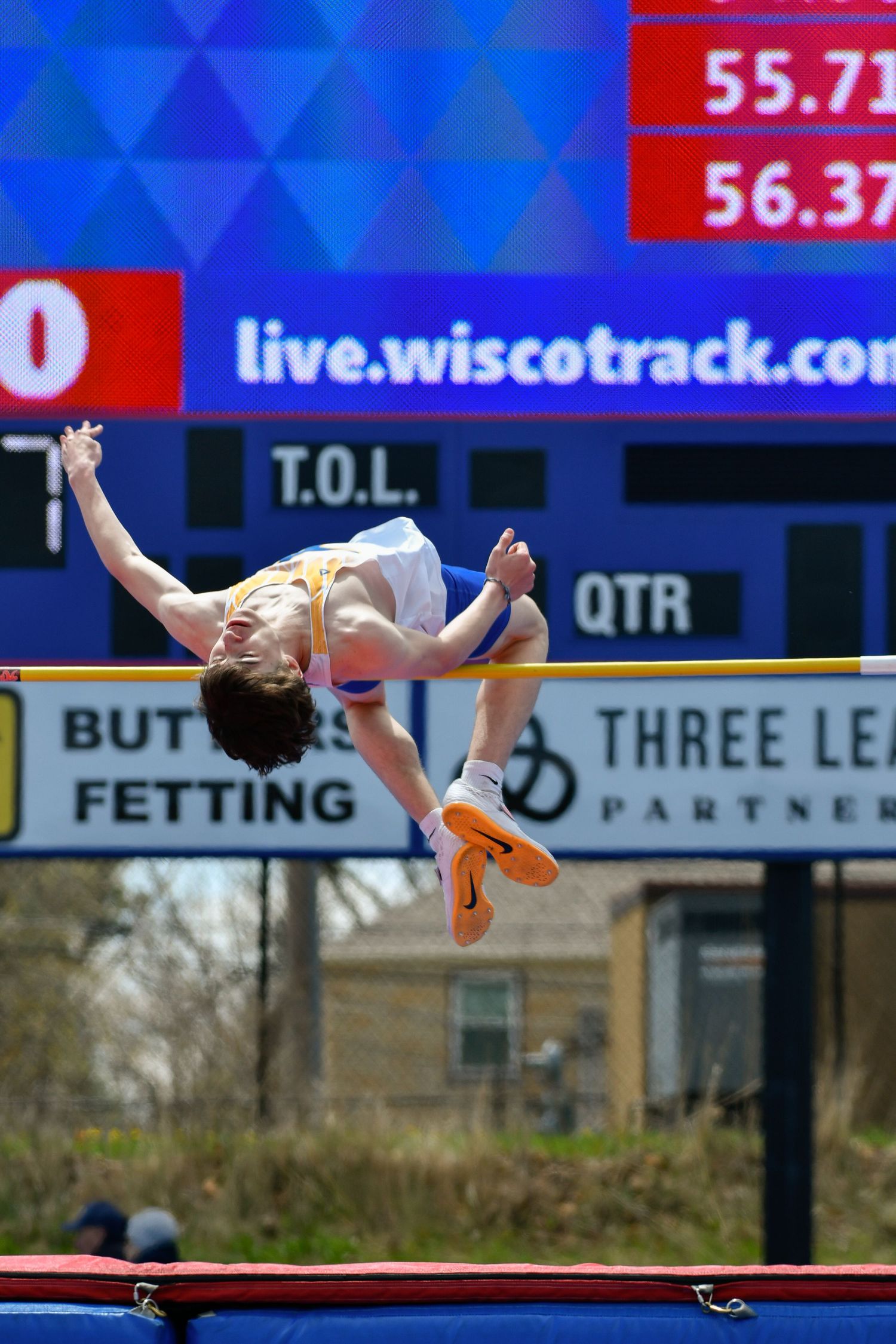 Athletes performing high jump at track meet with electronic scoreboard visible.