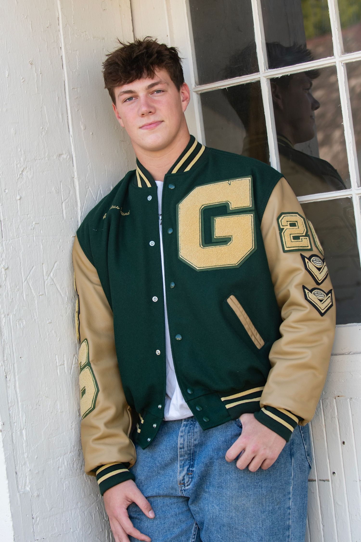 Student wearing a green and tan varsity letterman jacket with jeans poses against white wall panels.