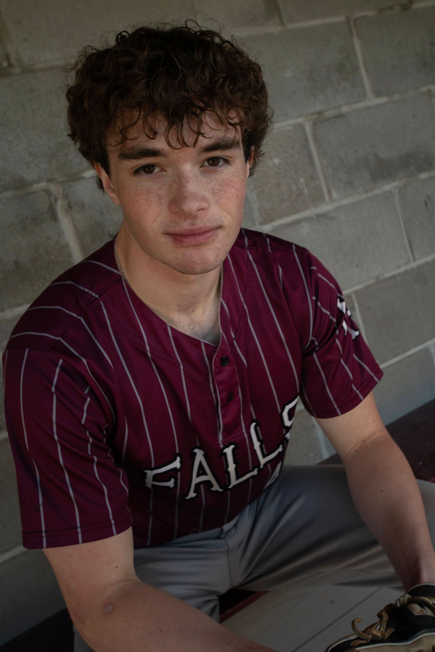 Young baseball player in maroon pinstripe Falls jersey sitting in dugout, curly hair, freckles, game day portrait.