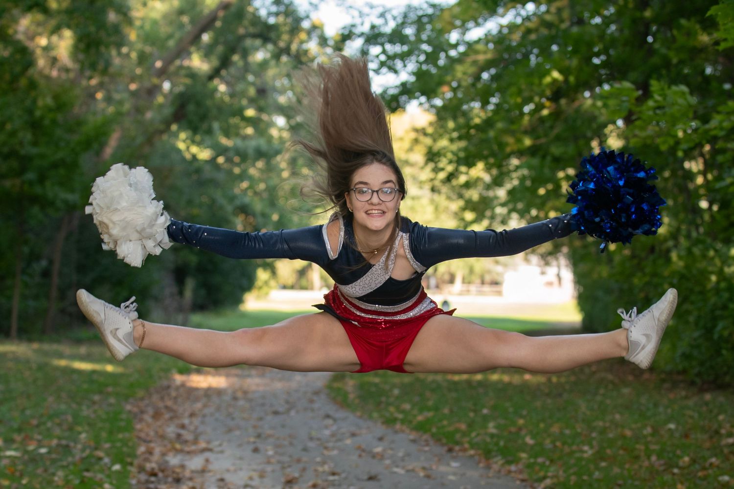 A cheerleader performs a split jump outdoors in a park setting with trees in the background.