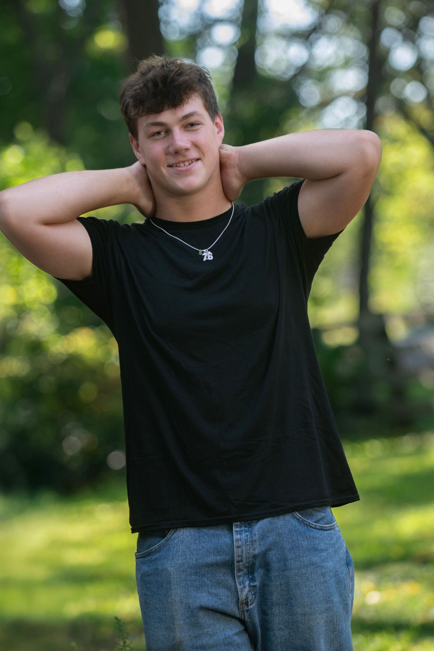 A person in a black t-shirt and jeans poses casually outdoors against a green natural background.