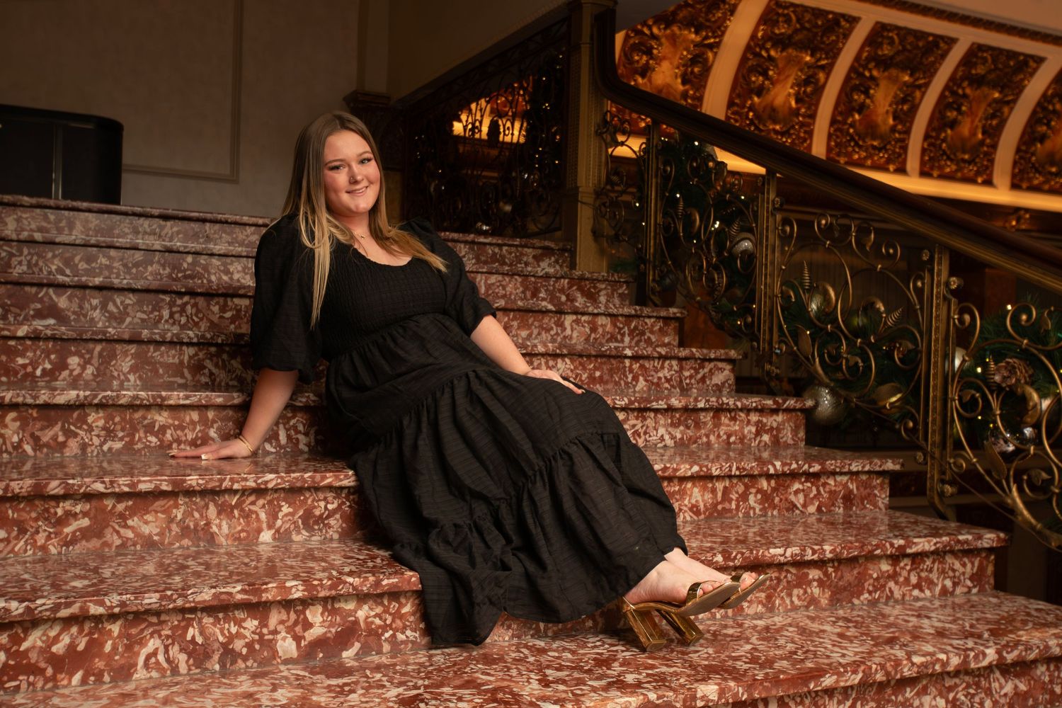 Woman in black dress poses elegantly on marble stairs with ornate gold railing in a grand venue.