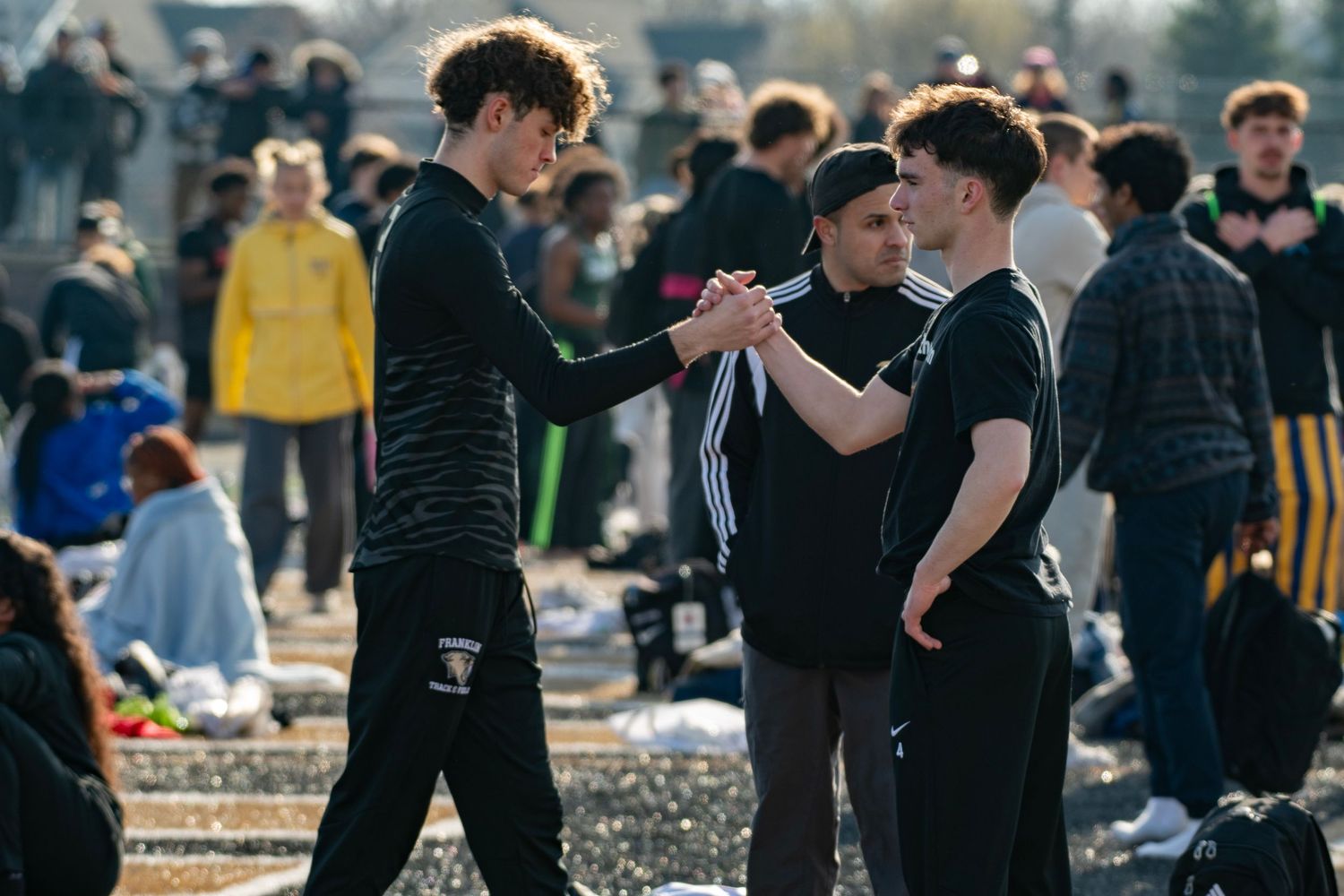 Two young men in black athletic wear shake hands at an outdoor track meet with spectators in the background.