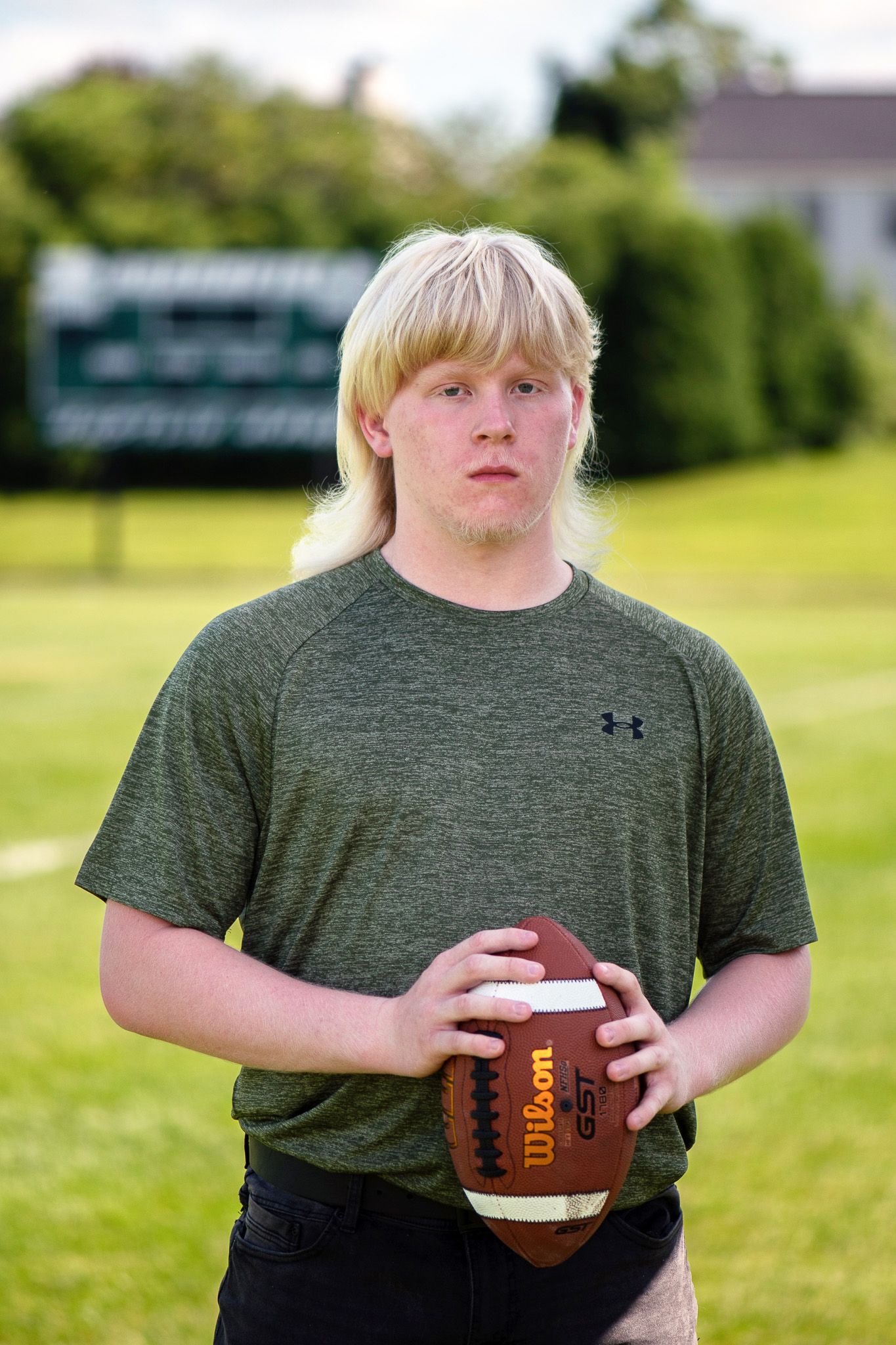 Someone in a green Under Armour shirt holds a football while standing on a grassy field. Game Day to Grad Day Senior Portrait Experience