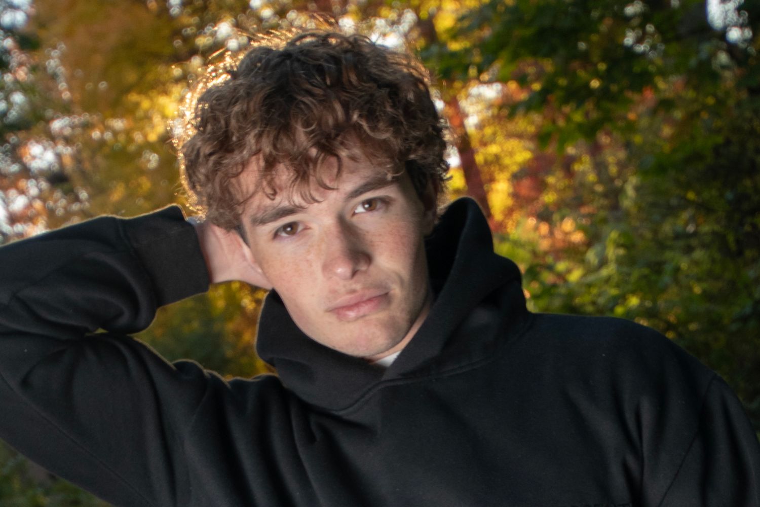 Young man with curly hair in black hoodie poses outdoors among colorful autumn foliage.