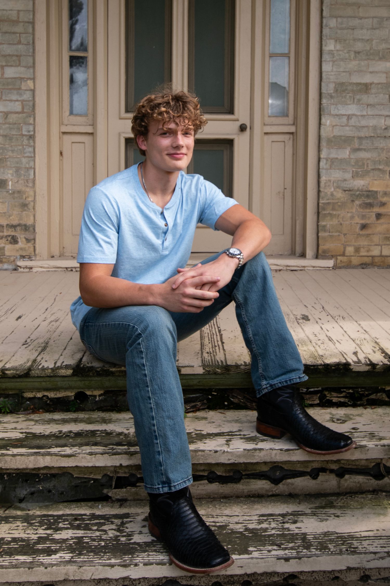 A young person in a light blue henley shirt and jeans sits casually on outdoor stone steps near a beige door.