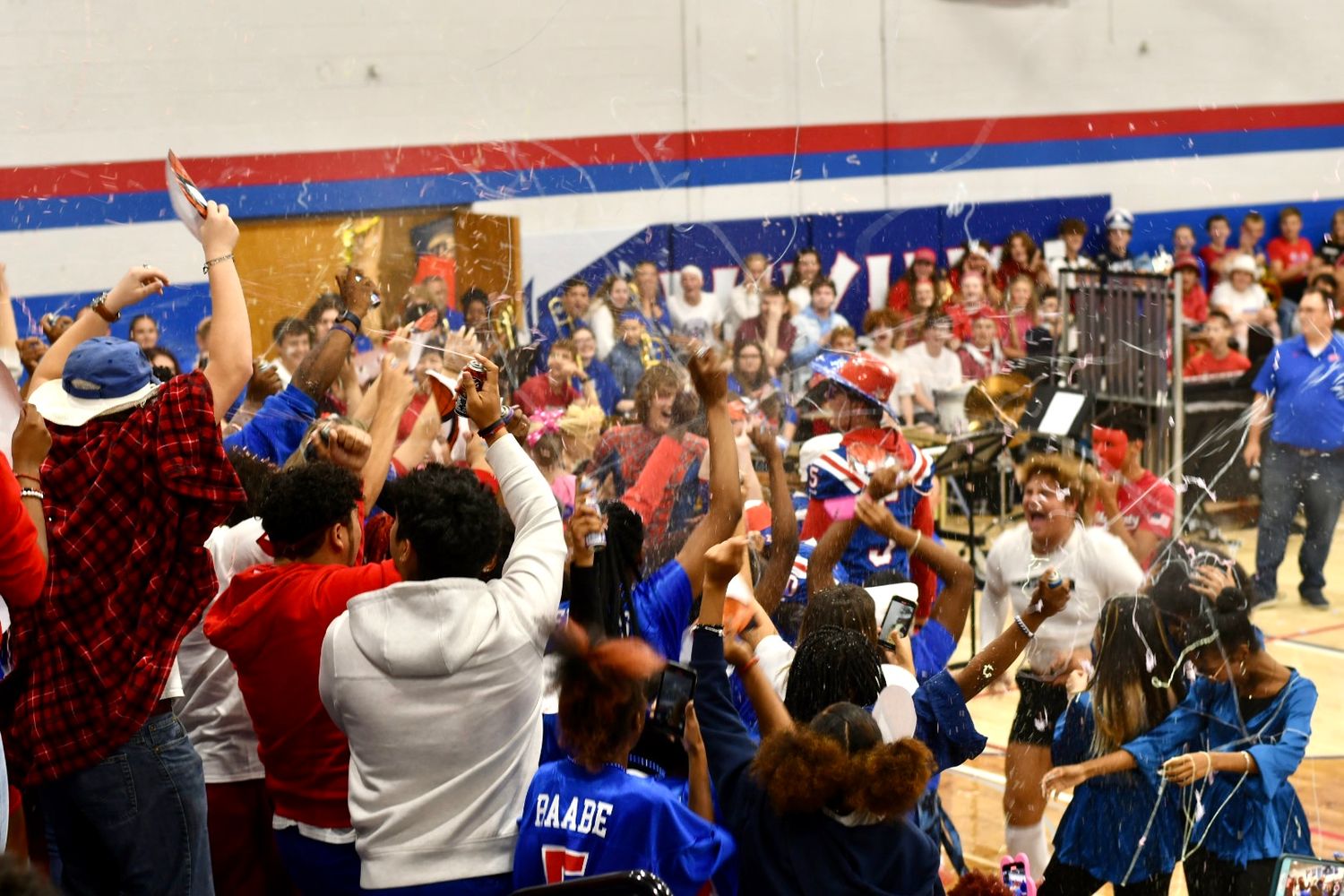 Students celebrate in a school gymnasium with confetti and raised hands during a lively pep rally event.