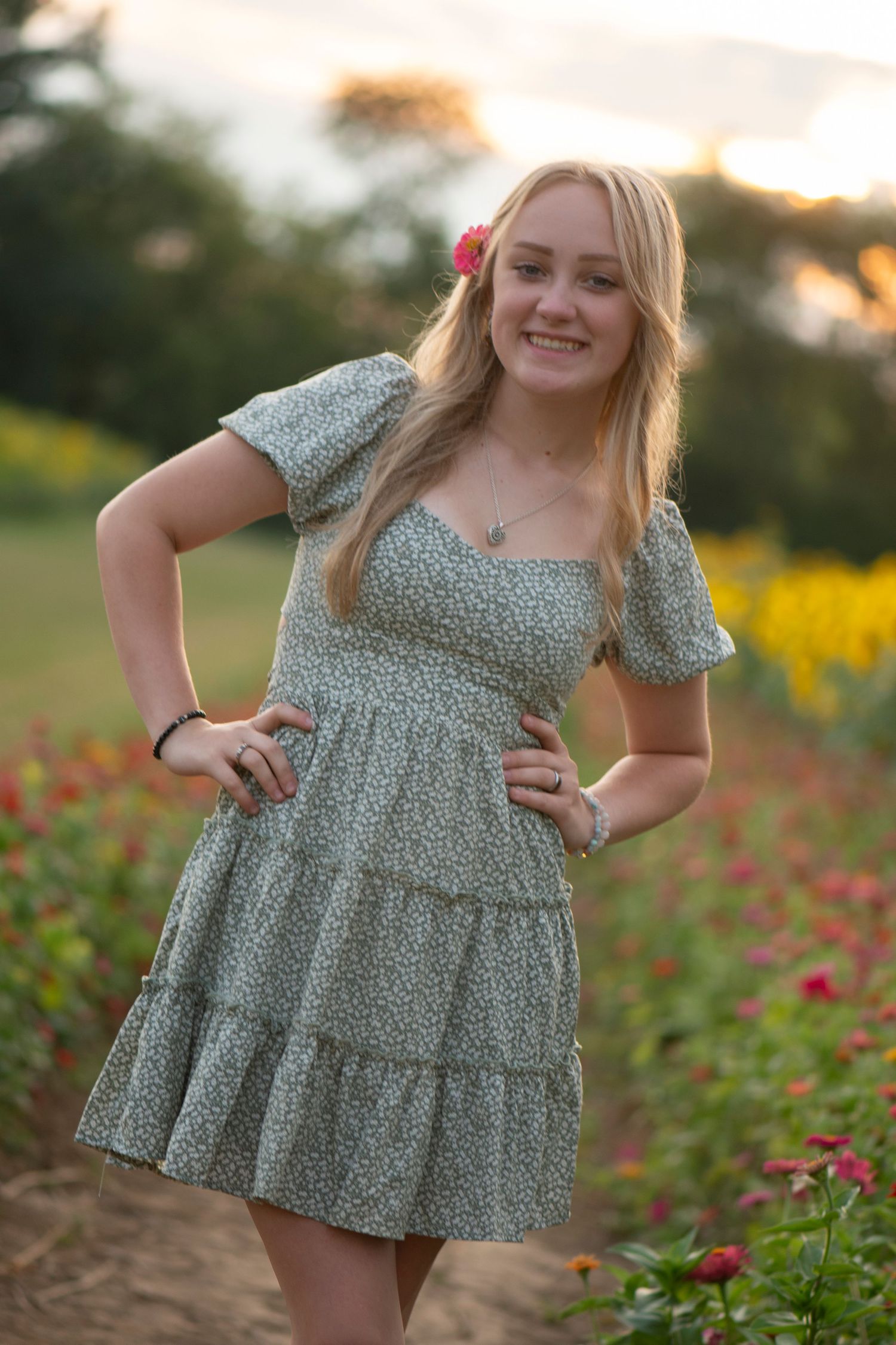 Young blonde woman in floral dress poses in colorful flower field at sunset with pink flower in hair.