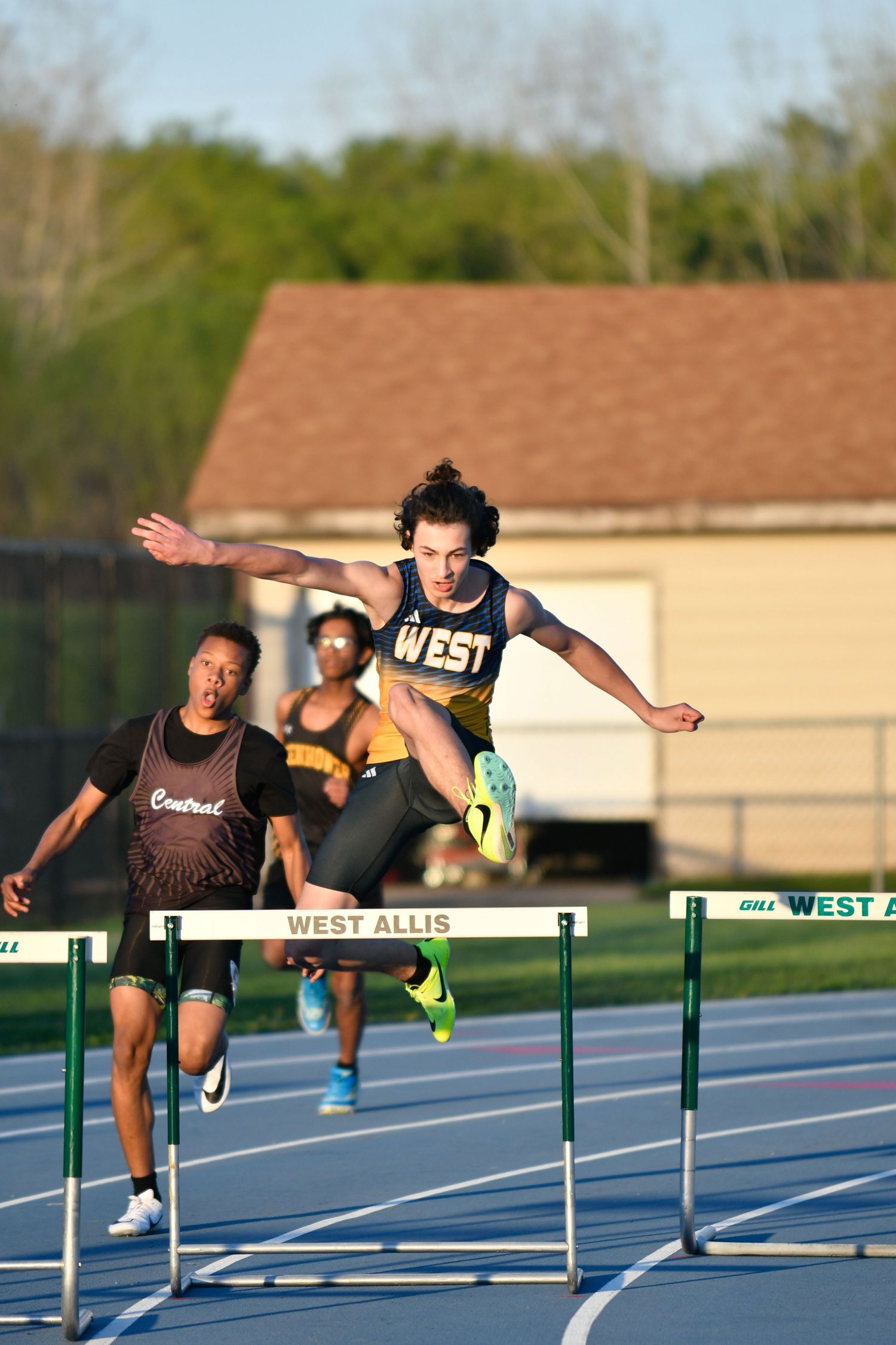 Hurdlers competing in a race on a blue track during sunset.