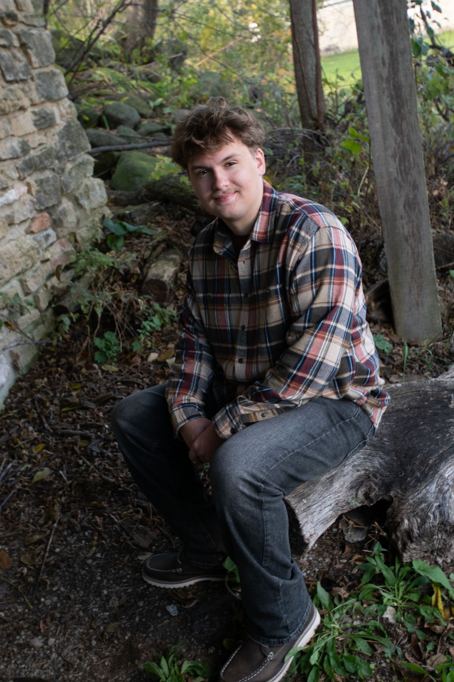 Young man in plaid shirt sitting on log near rustic stone wall outdoors.