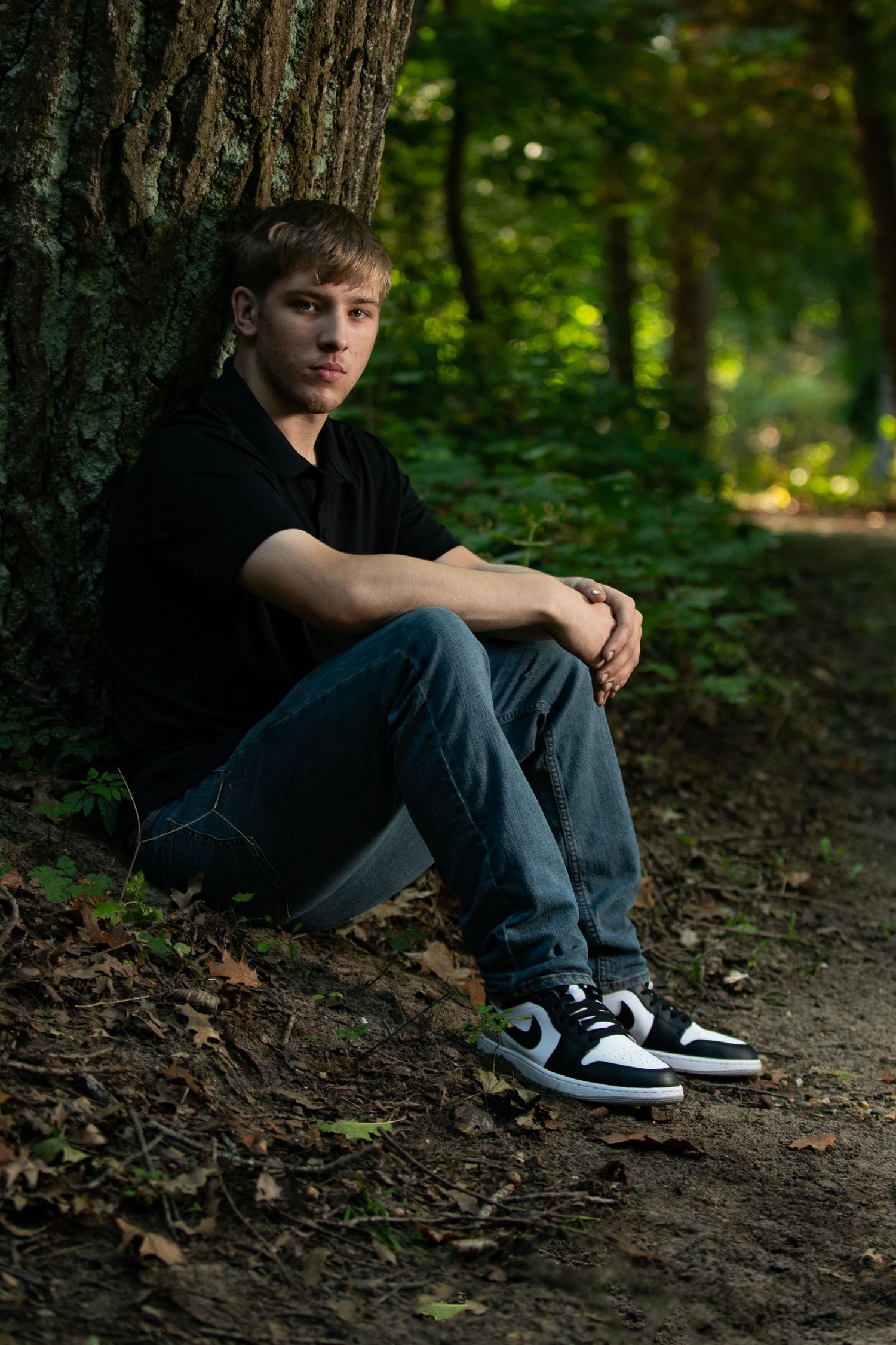 Portrait in a forest setting showing someone sitting against a tree trunk wearing black Nike sneakers and casual attire.