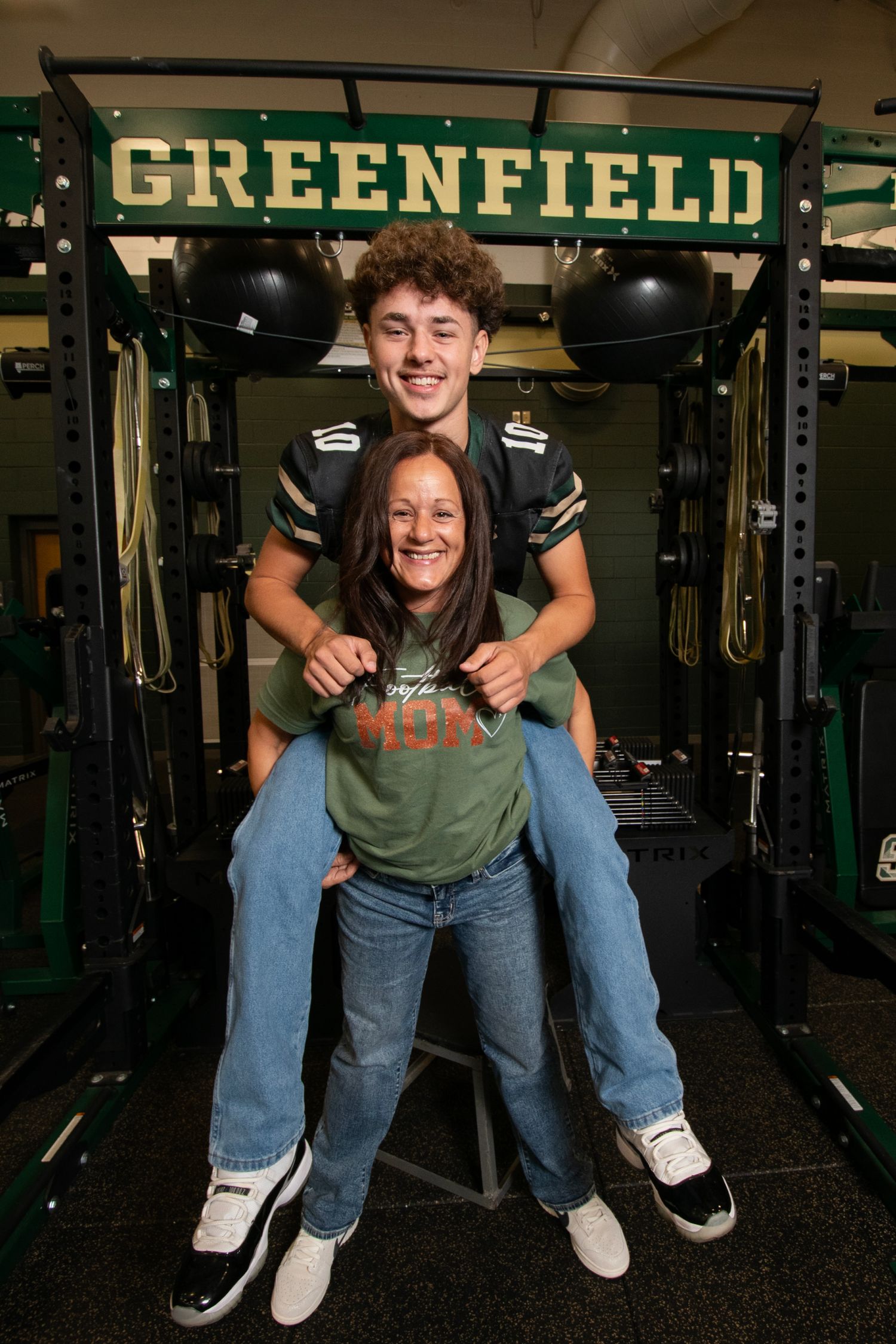 Two students pose playfully in a school weight room with 'GREENFIELD' signage and exercise equipment visible in background.