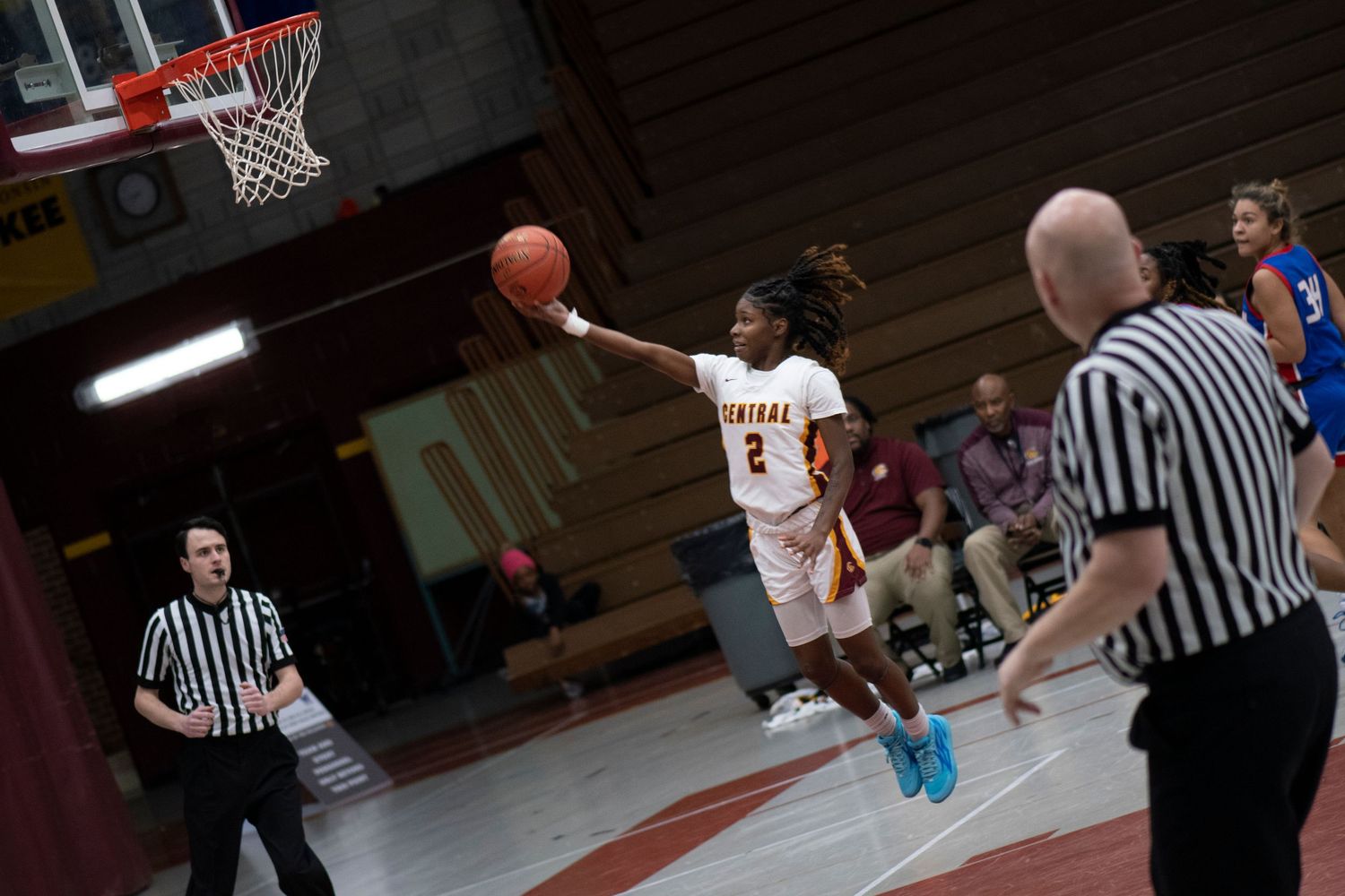 Female basketball player #2 in white uniform leaps toward hoop during indoor game with referees watching.