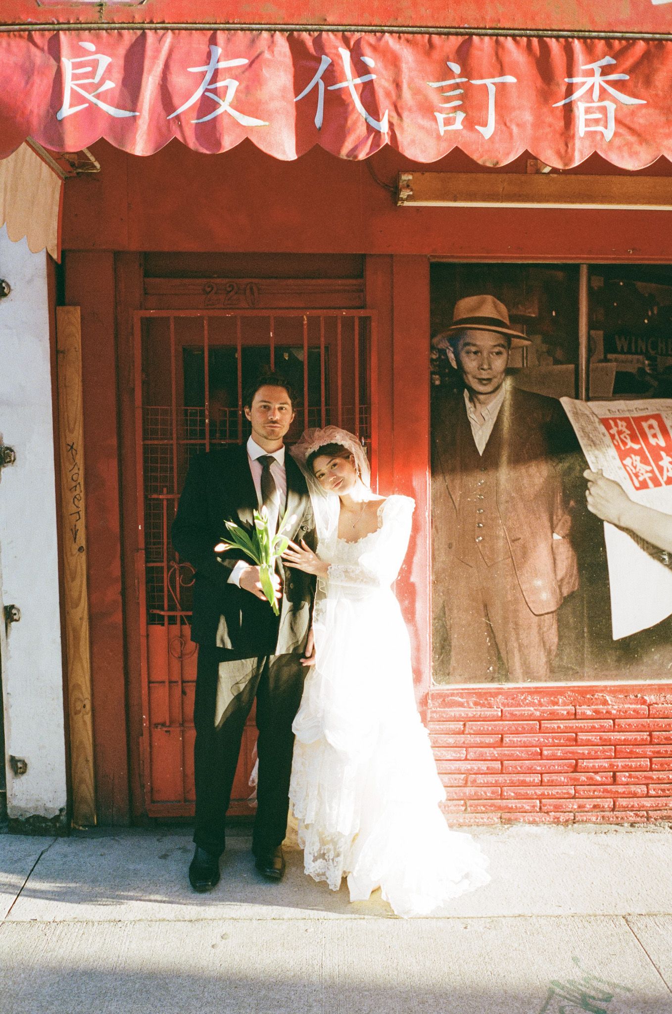 A couple in formal wedding attire poses in front of a traditional red brick building with Chinese signage.