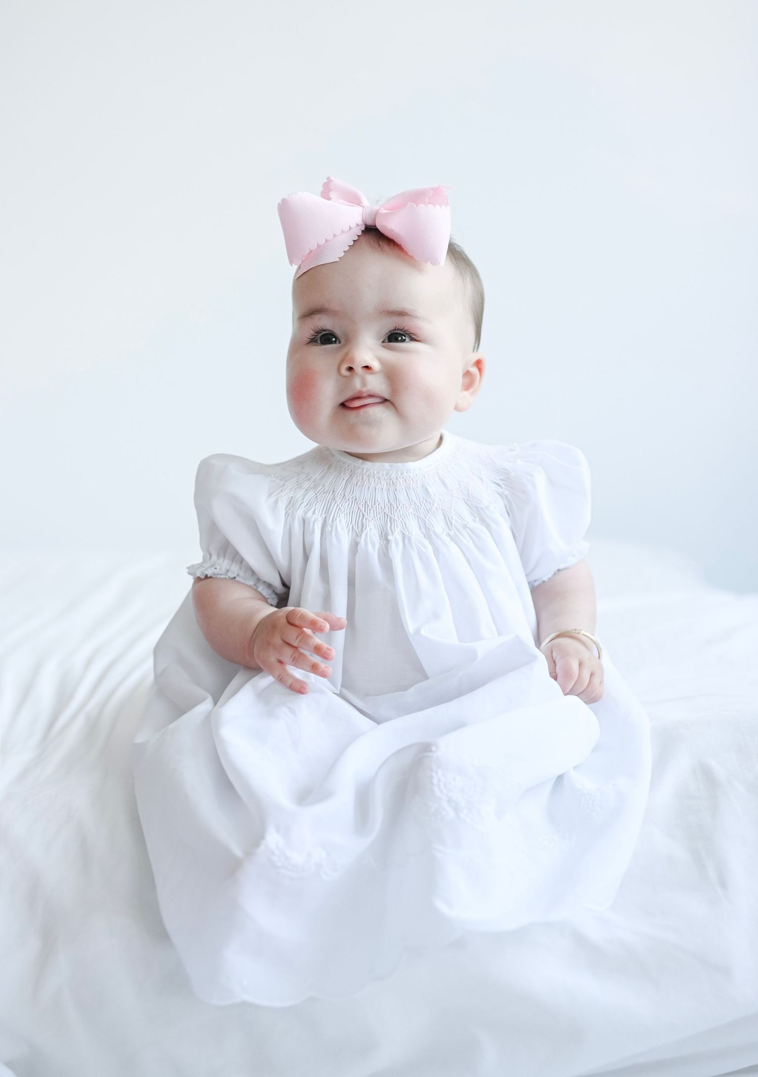 Baby in flowing white dress and pink bow against light blue background.