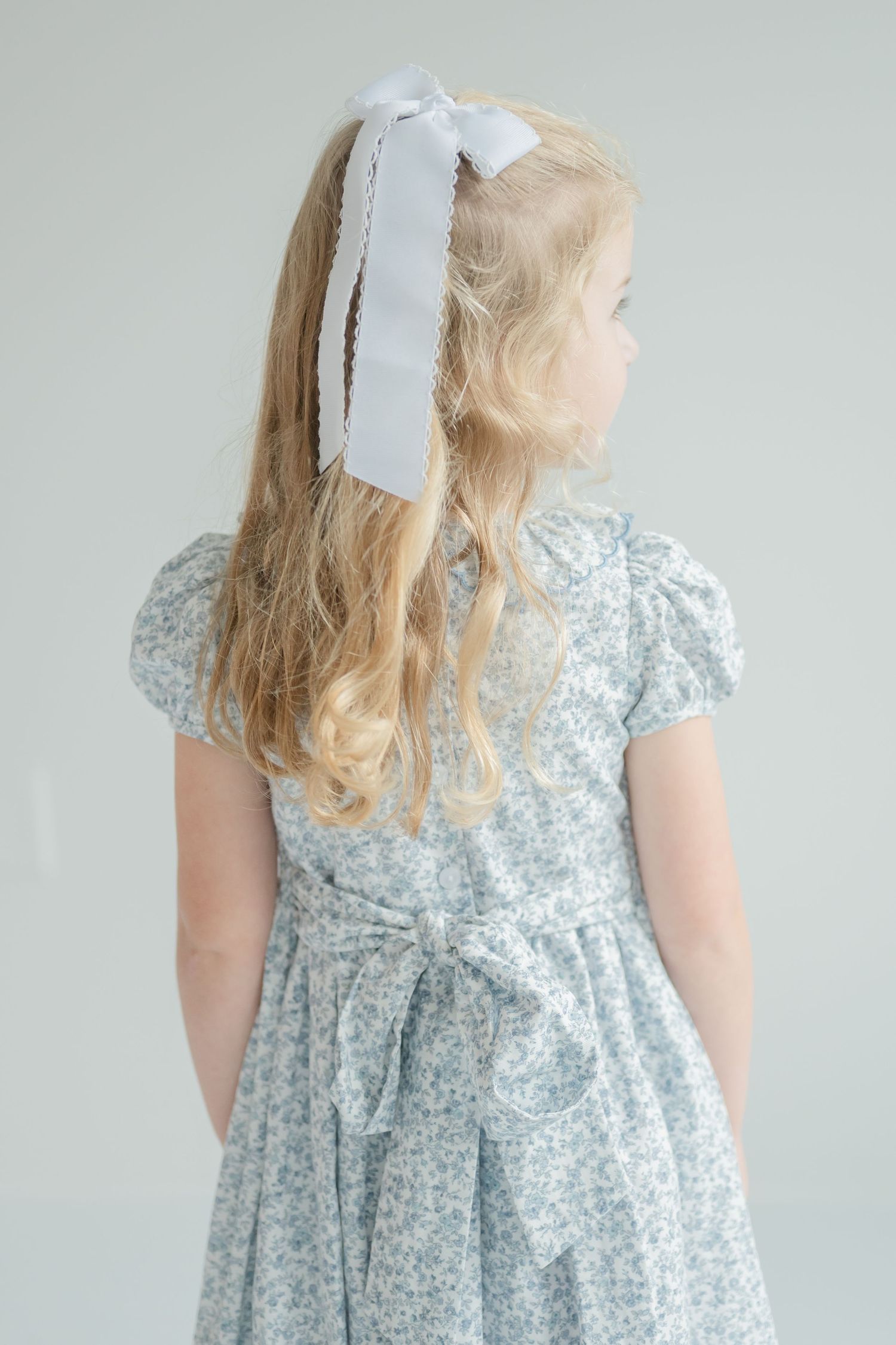 Studio portrait of a young girl with long blonde curls down her back, in a floral blue dress and white hair ribbon, looking away.