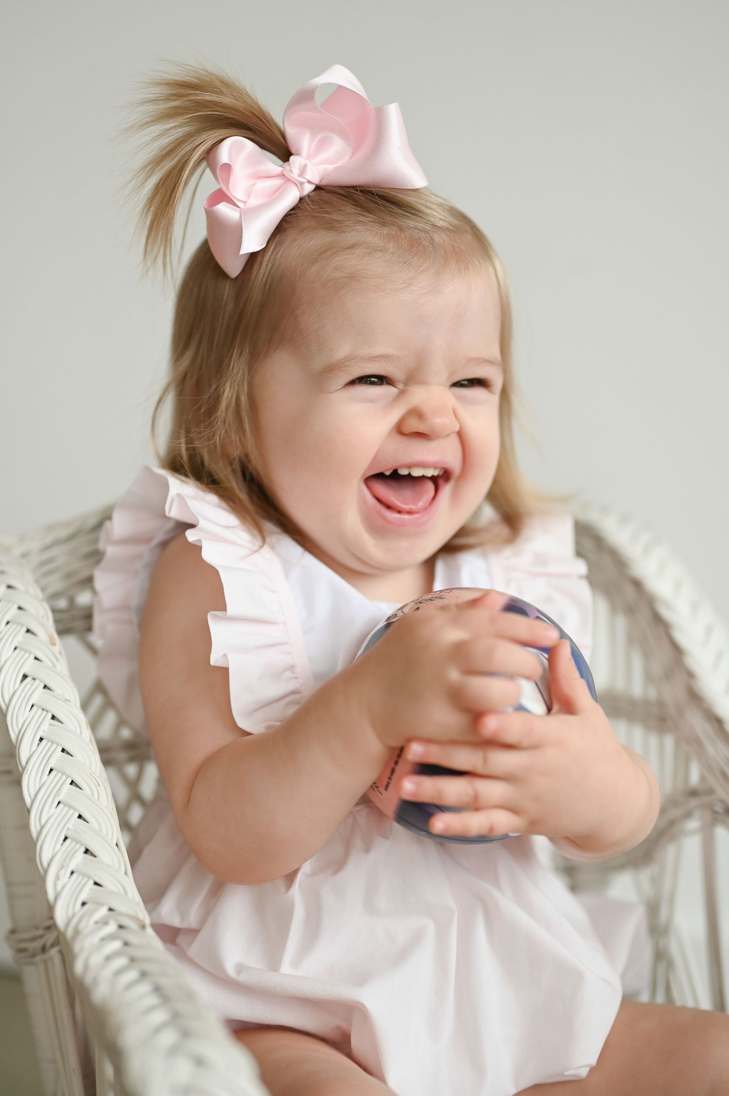 Baby in white ruffled dress and pink bow sitting in wicker chair expressing joyful laughter.