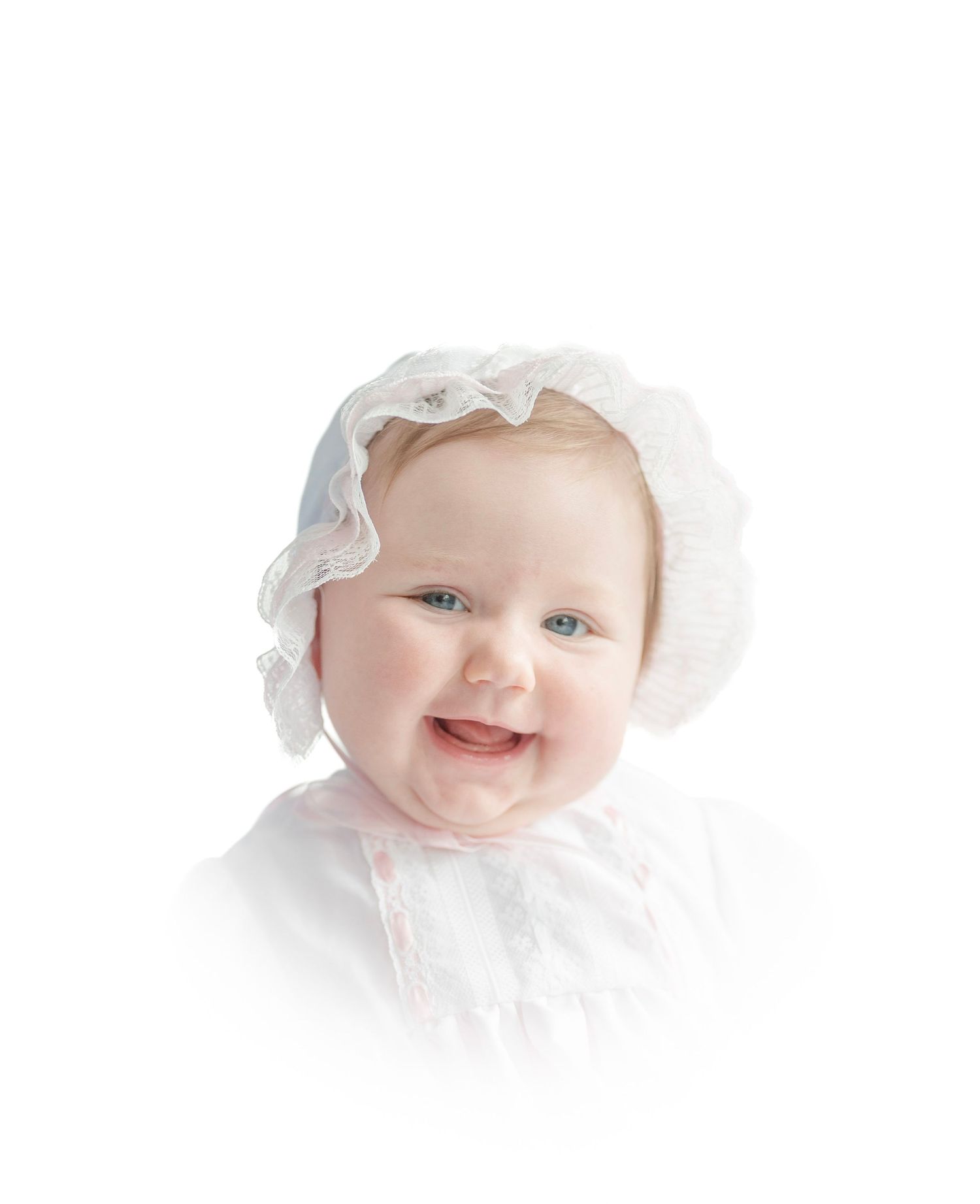Portrait of a smiling baby wearing a ruffly white bonnet and christening gown against a white background.