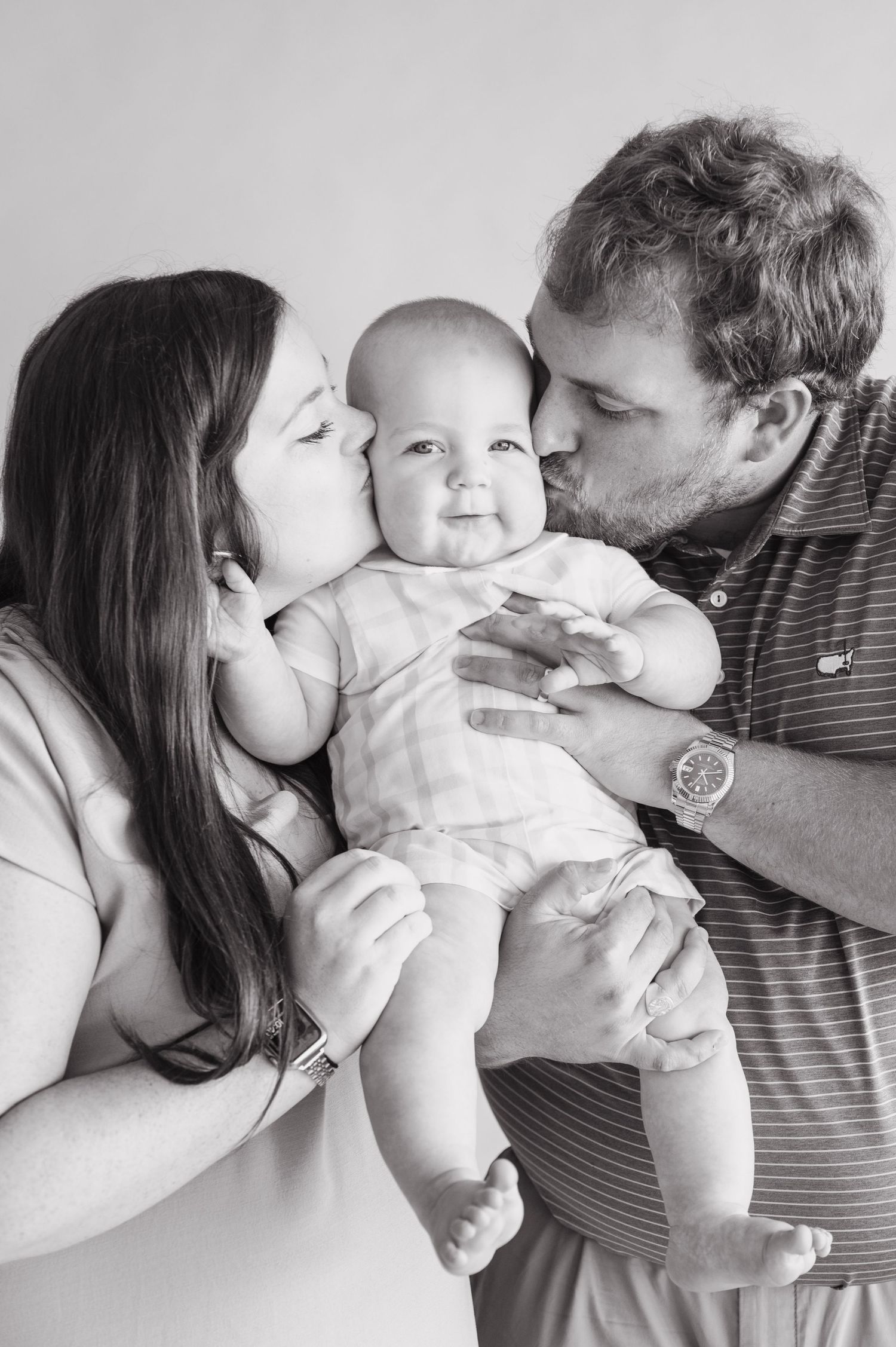 Black and white portrait of parents kissing baby's cheeks in studio setting.