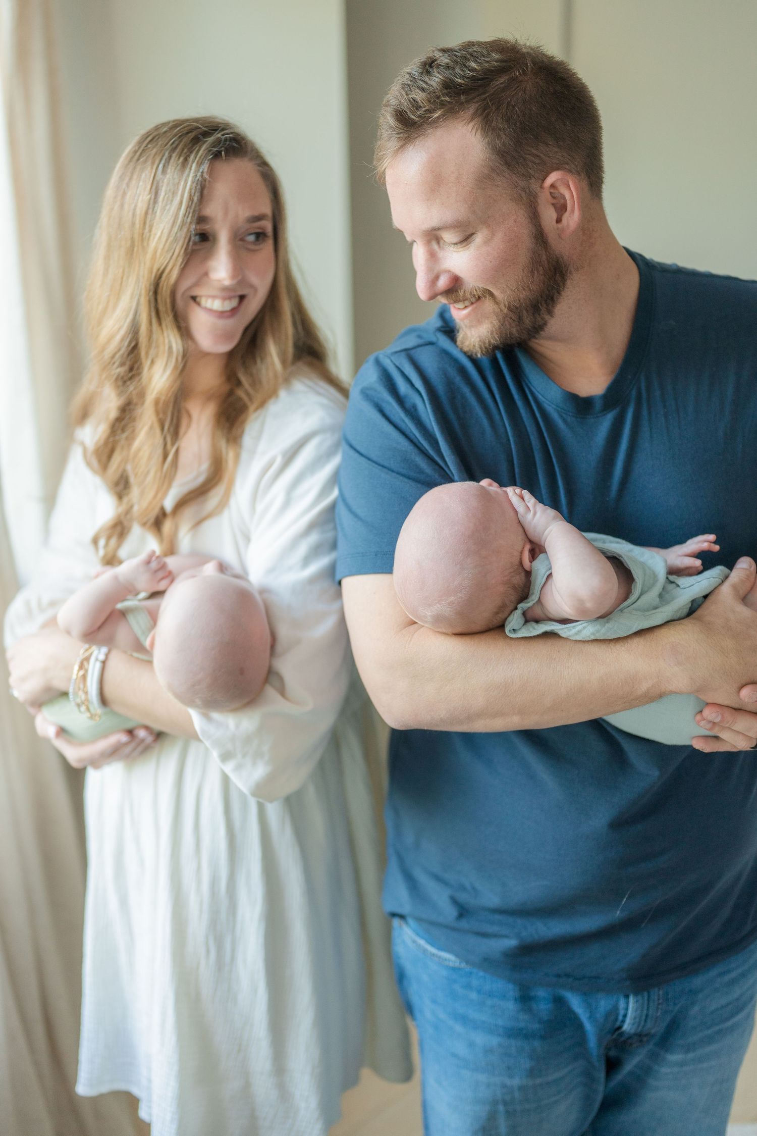 A young woman and man in casual clothes hold their twin babies in the light of a nearby window.