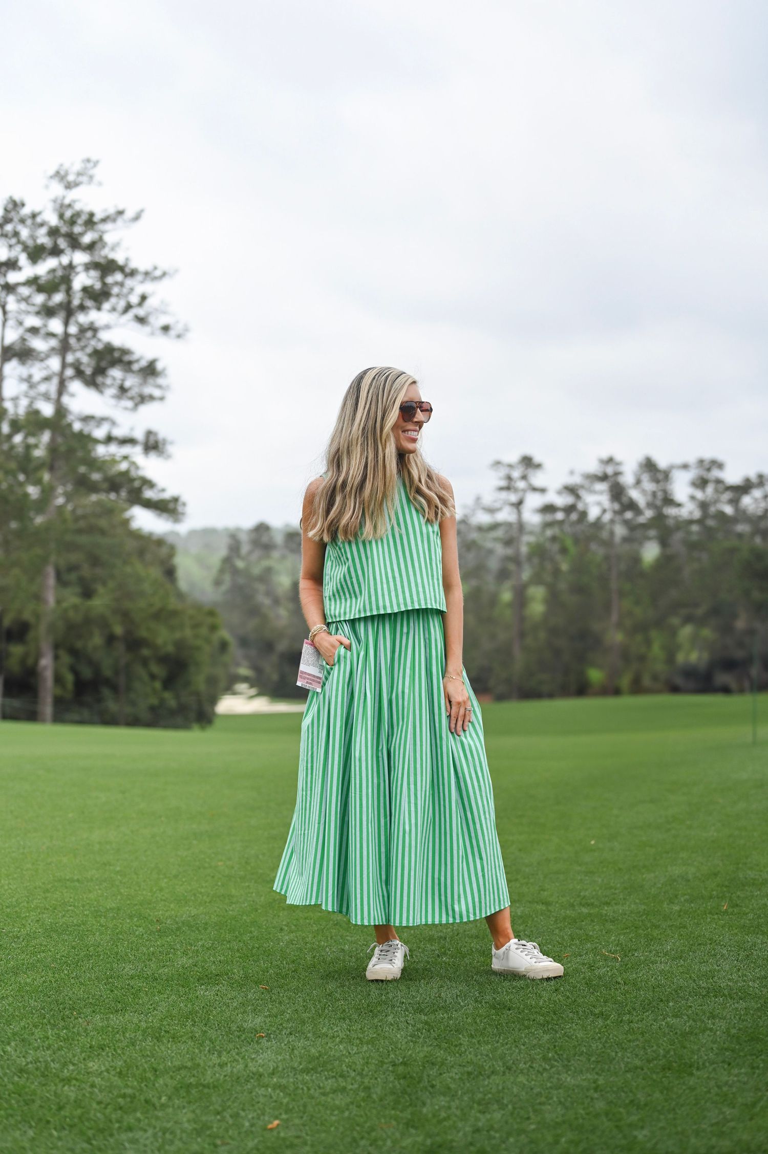 A young woman with wavy blonde hair and sunglasses, wearing a flowing green and white striped sundress on a lush golf course.