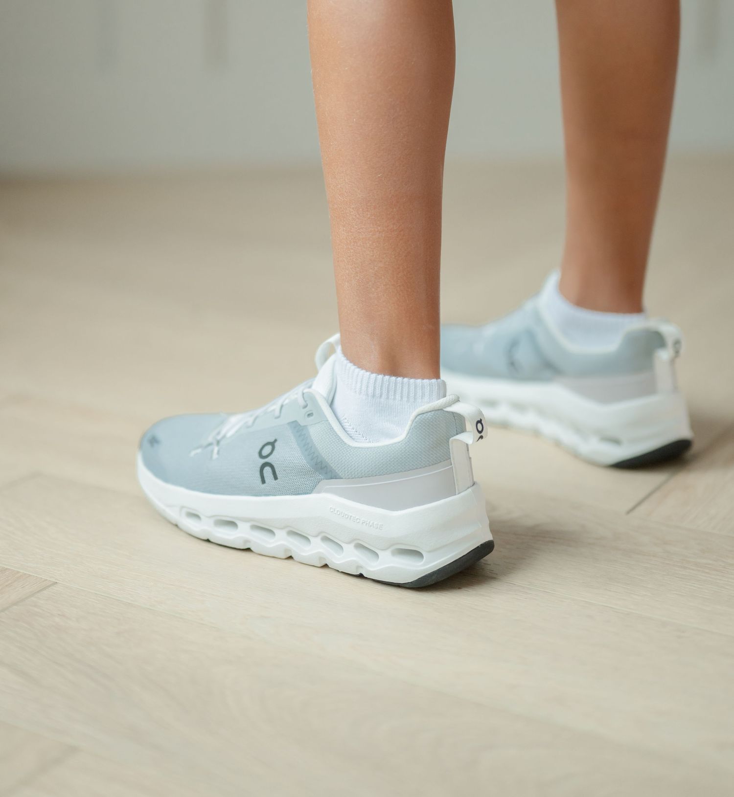 Low closeup of white On Cloud running shoes worn with white ankle socks, seen from behind and to the left, showing the heels and sides.