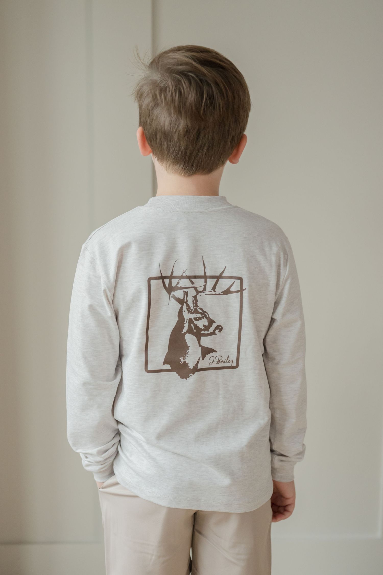 Studio shot, back view of a young boy with brown hair in khakis and a gray sweatshirt with a deer logo.