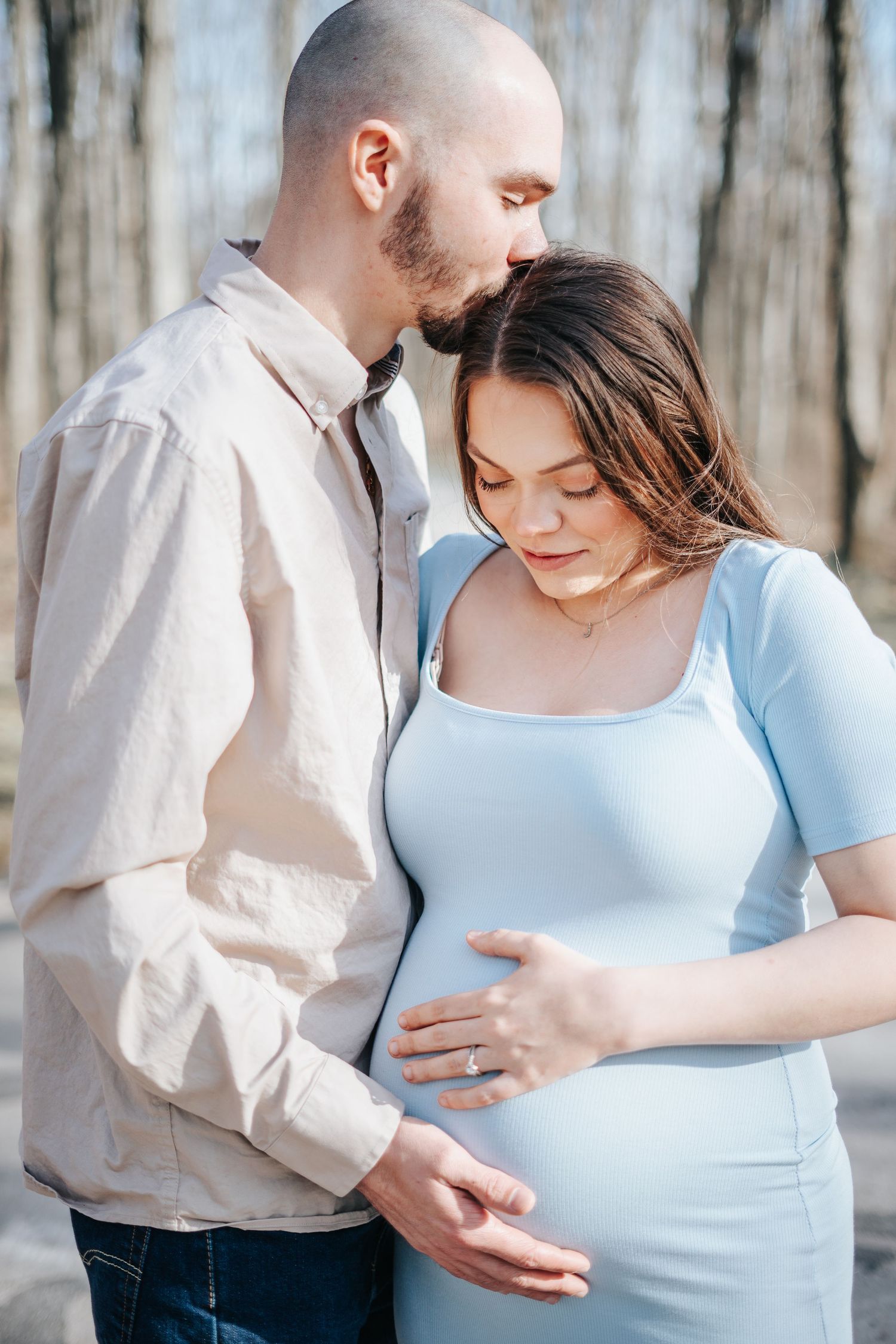 A couple shares a tender moment outdoors, with the man kissing his pregnant partner's head as she cradles her baby bump.