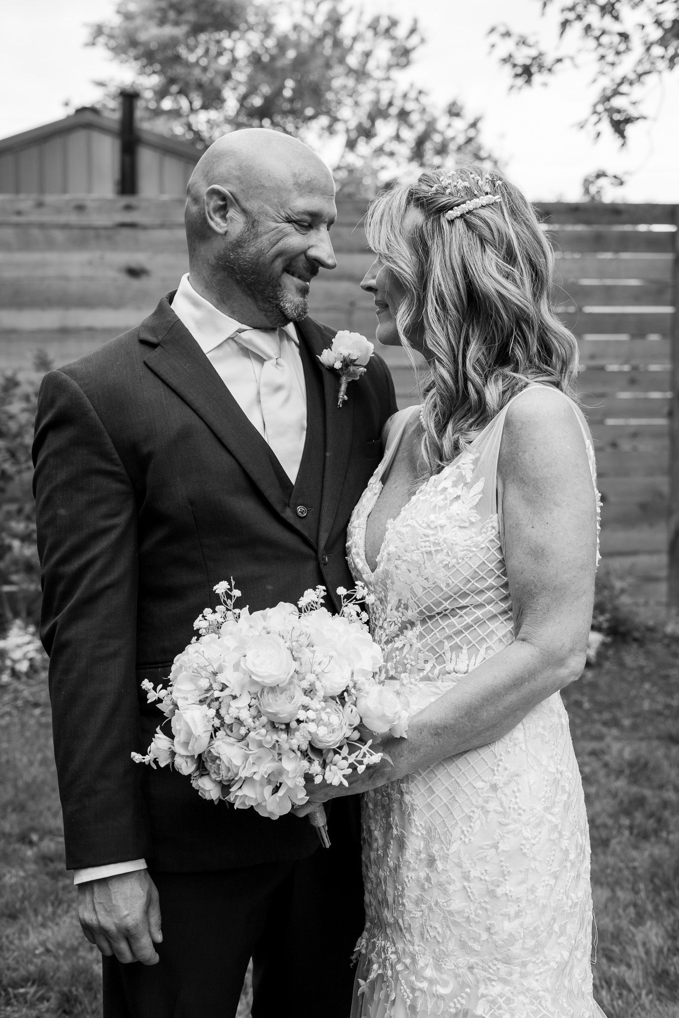 Black and white photo of a smiling bride and groom sharing a tender moment outdoors, she holds a bouquet of roses and wears a lace gown.