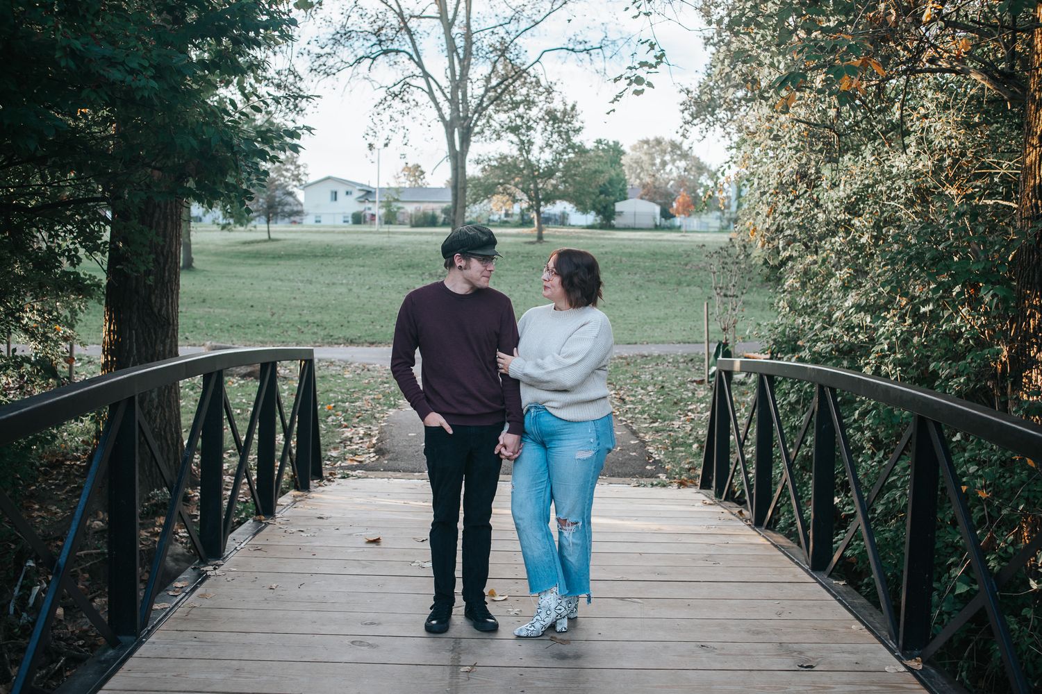 Couple standing close together on a wooden footbridge with black railings, surrounded by autumn trees and a green park with houses behind.