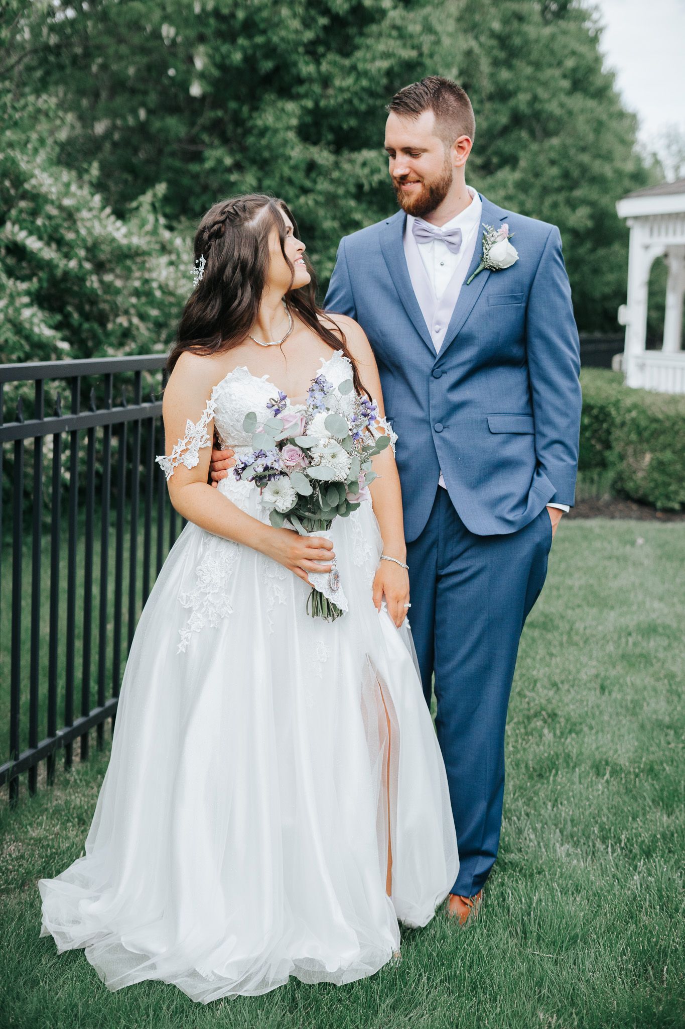 A bride in a white tulle gown holding a pastel bouquet gazes up at her groom in a blue suit and bow tie on a green lawn.