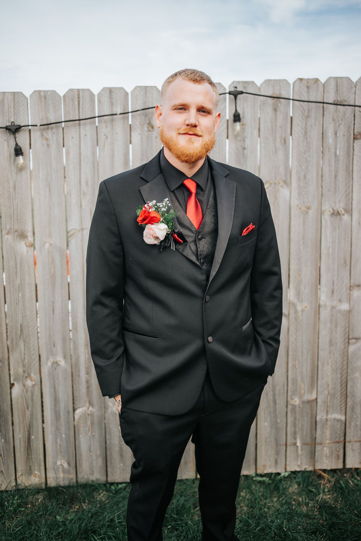A groom in a black suit with red tie and boutonniere poses against a wooden fence on his wedding day.