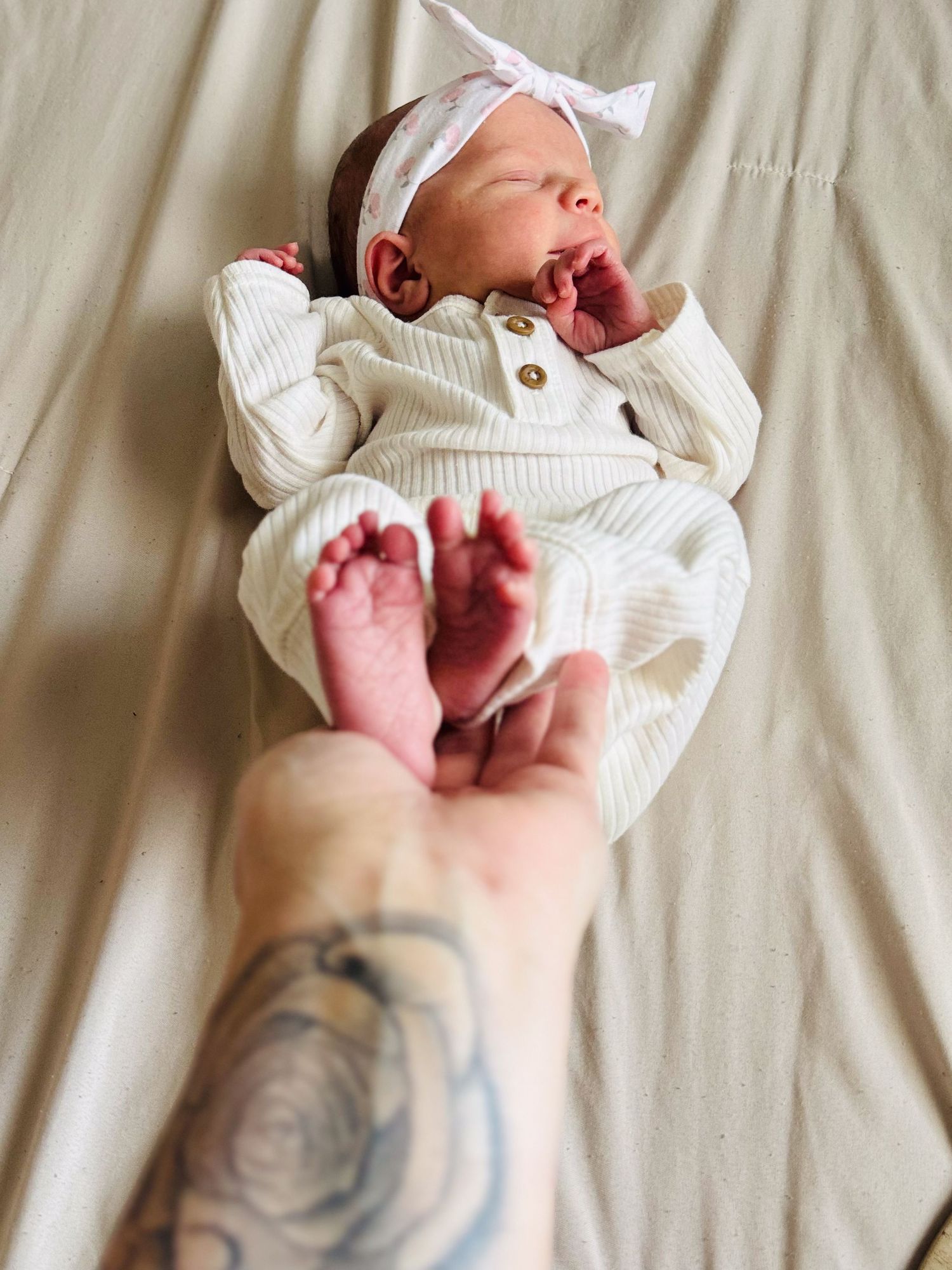 Tattooed parent holding newborn baby's tiny feet, infant in white ribbed outfit and pink bow headband lying on beige blanket.