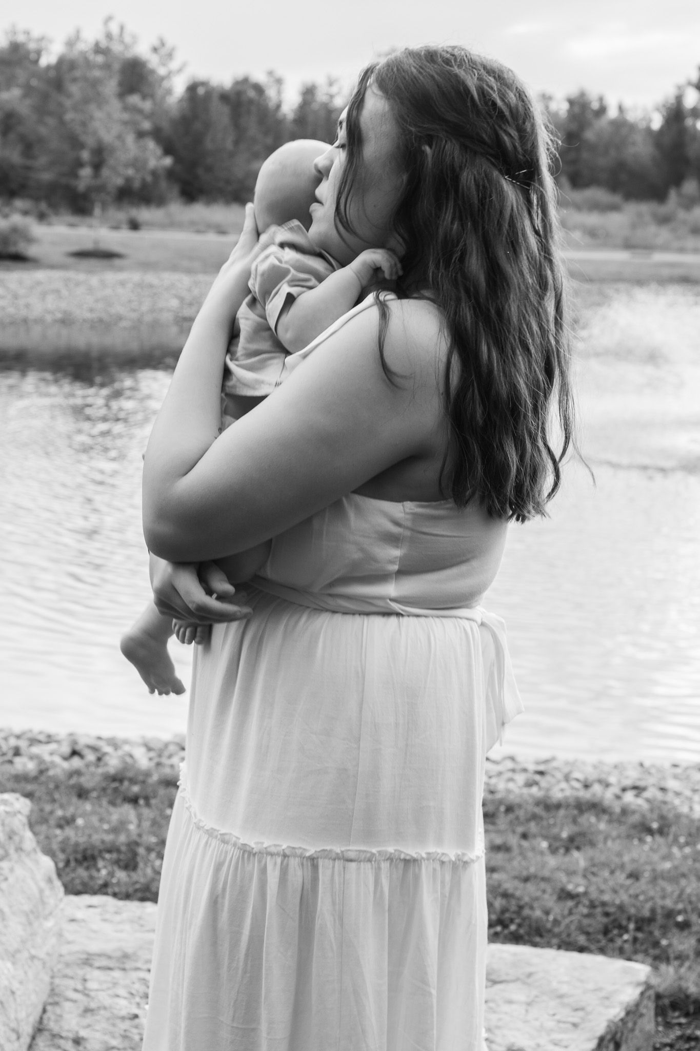 Black and white photo of a mother holding her newborn baby by a serene lake, wearing a flowing white dress.