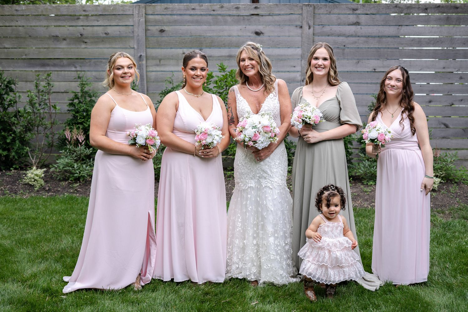 Wedding party wearing pink bridesmaid dresses and a white bridal gown pose together on grass in front of wooden wall.