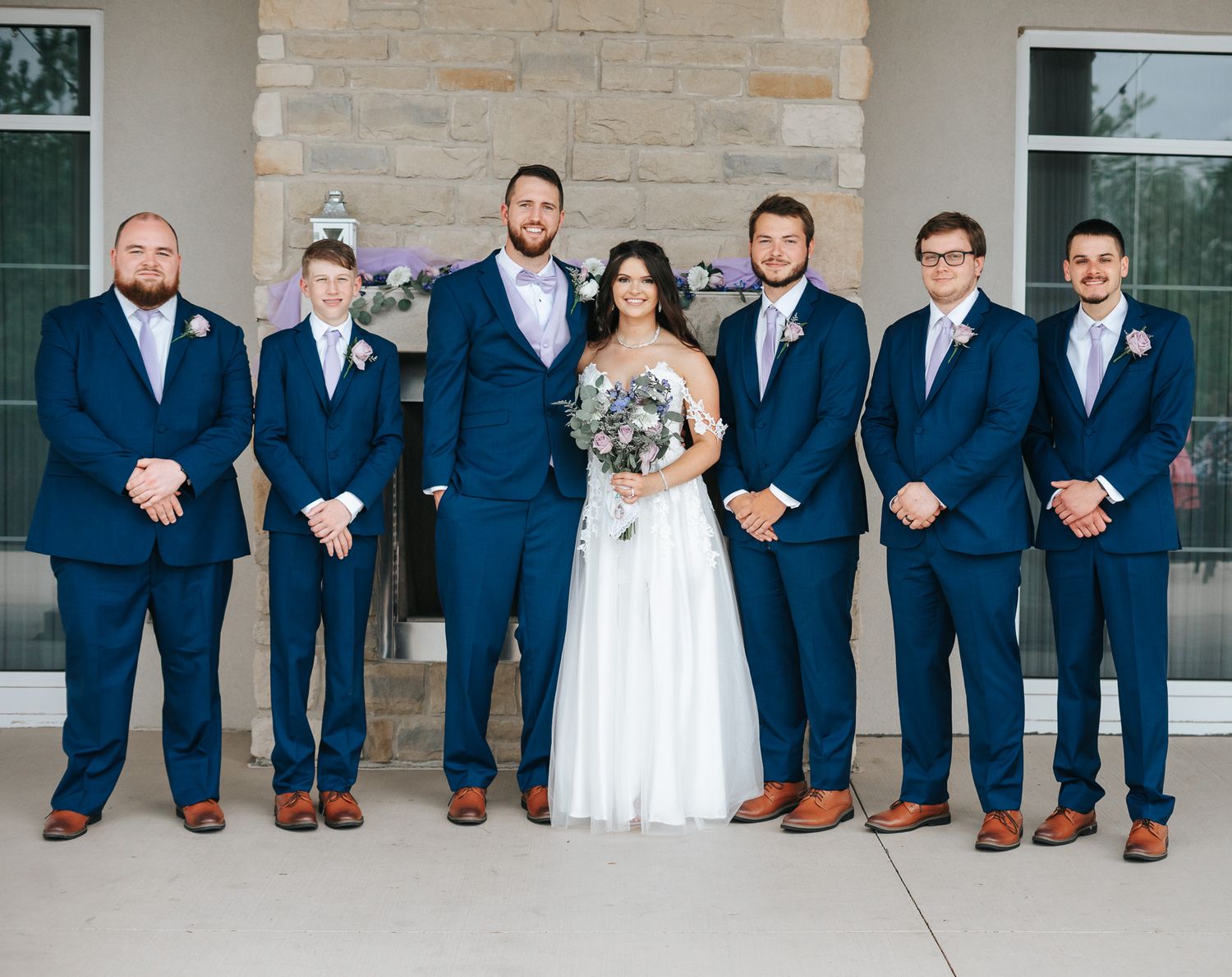 Bride in a white gown holding a bouquet stands among six groomsmen in navy suits and brown shoes in front of a stone wall.