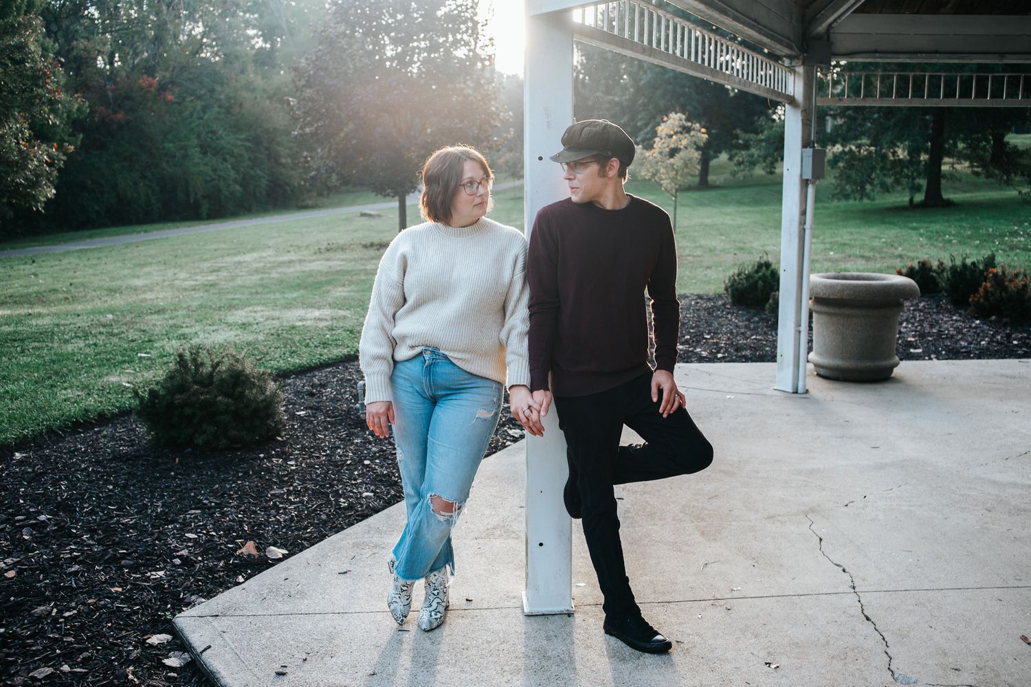 Two people hold hands while leaning against a white post on a sunlit covered patio overlooking a green lawn and trees.