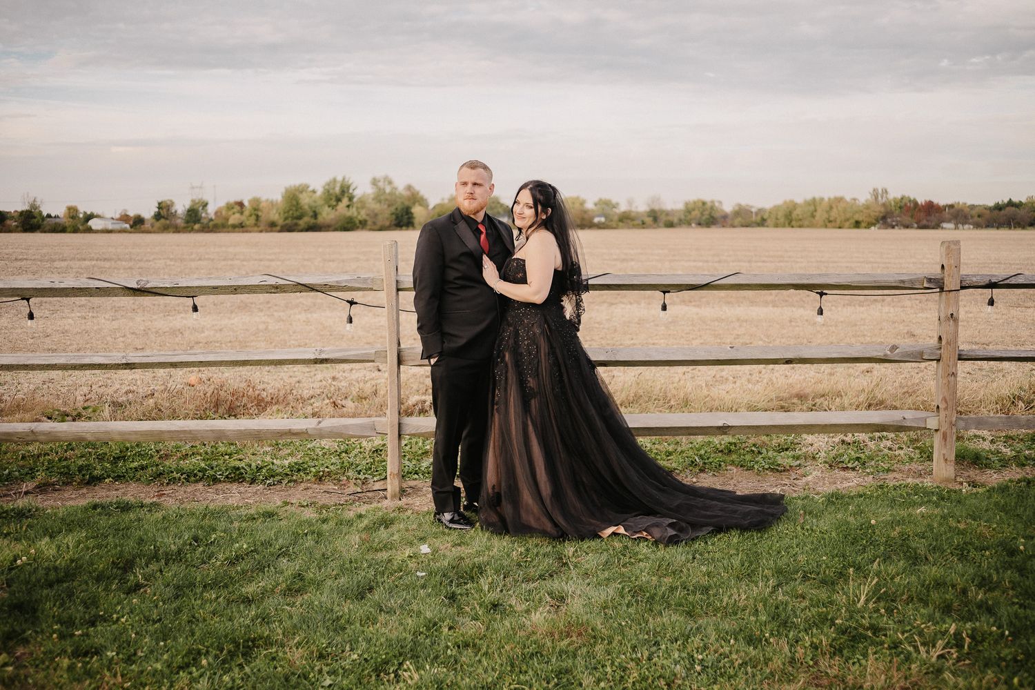 Couple poses in black wedding attire before a rustic wooden fence and open field at their gothic outdoor wedding.