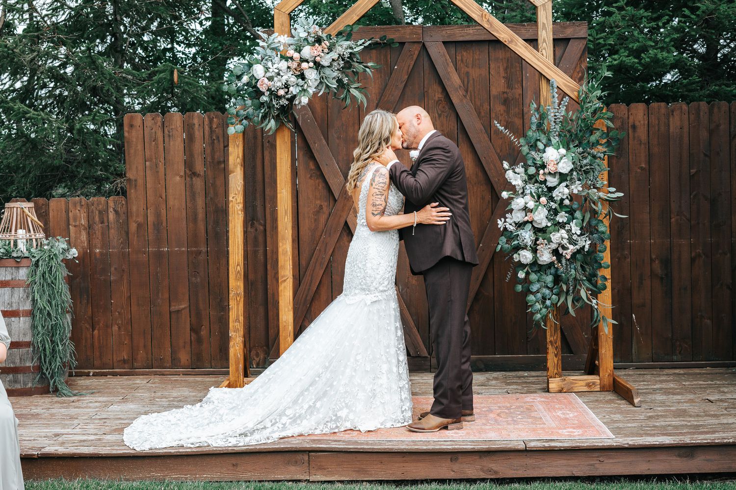 A romantic couple shares their first kiss under a rustic wooden archway decorated with white floral arrangements at an outdoor wedding.