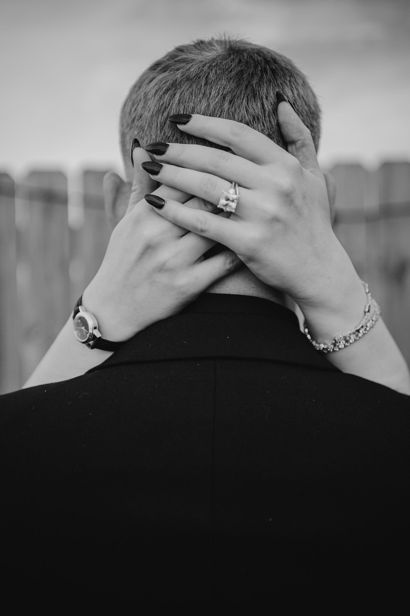 Black and white photo of a woman's hands with ring and bracelet embracing a man's head from behind.