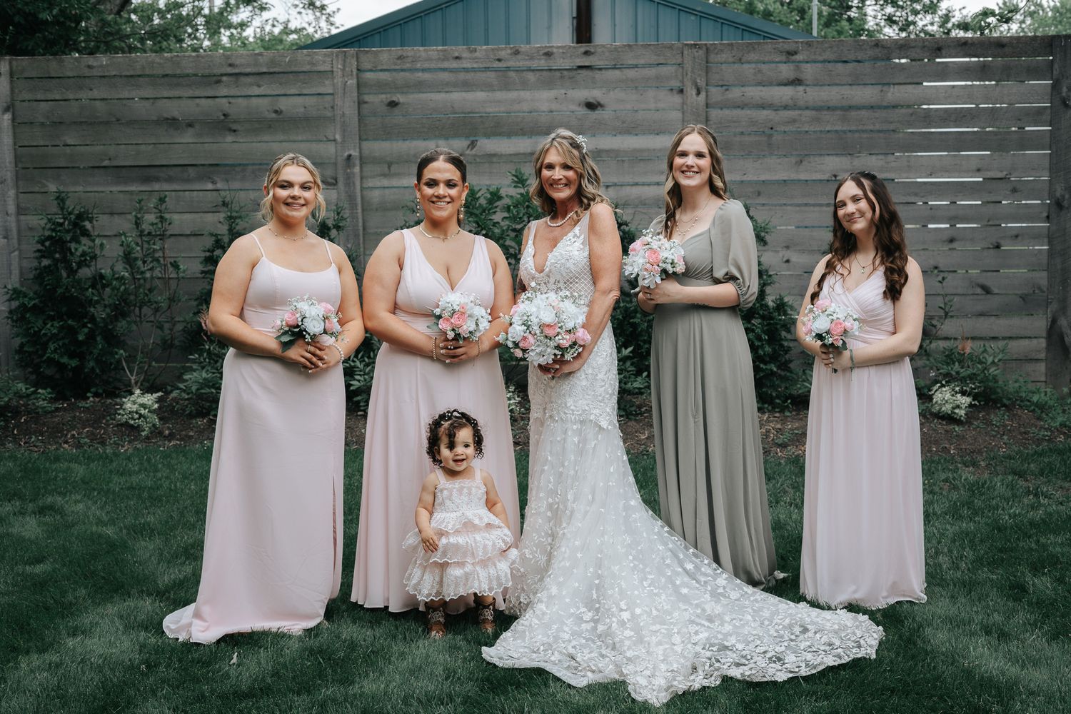 Bride in a lace wedding gown holding a bouquet with four bridesmaids and a toddler flower girl posing on a lawn by a wooden fence.