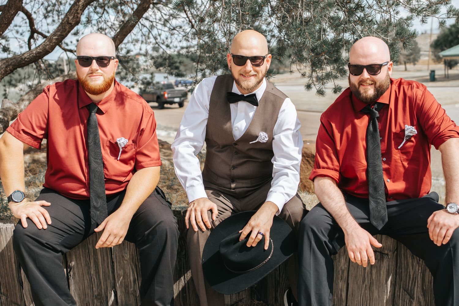 Groomsmen wearing sunglasses and red shirts pose together on wooden steps outdoors.