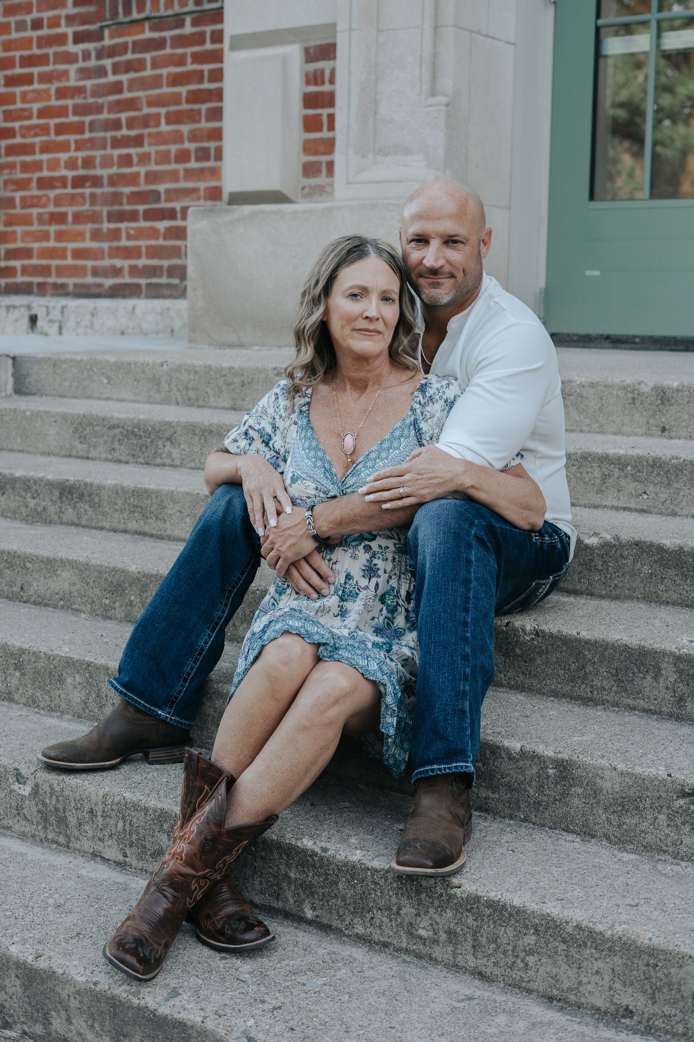 Couple sitting together on concrete steps by brick building wearing boots and casual attire.
